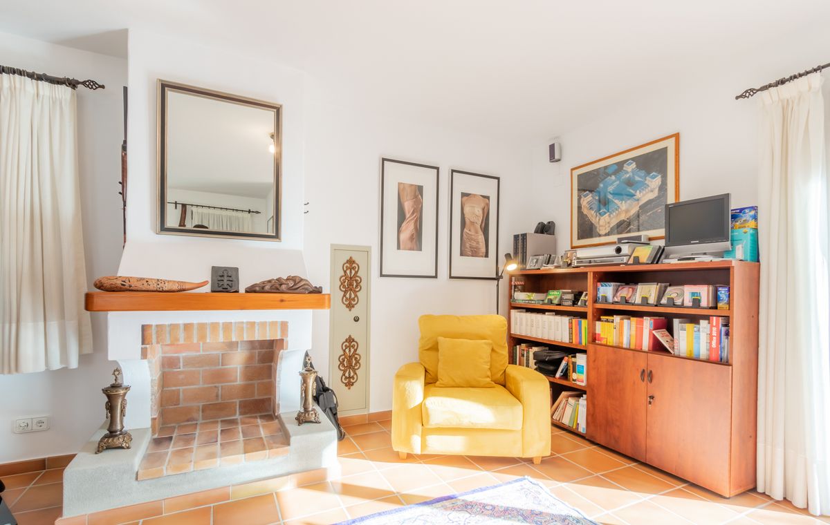 Woonkamer met een traditionele bakstenen open haard, een gele fauteuil en een houten boekenkast op een terracotta tegelvloer.