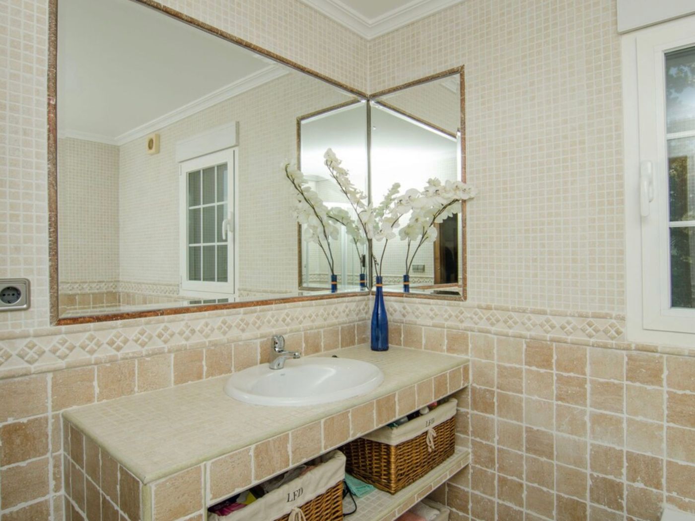 Badkamer met een betegeld wastafelblad, ingebouwde wasbak en open schappen met rieten manden. Grote hoekspiegels reflecteren de beige mozaïekmuren.