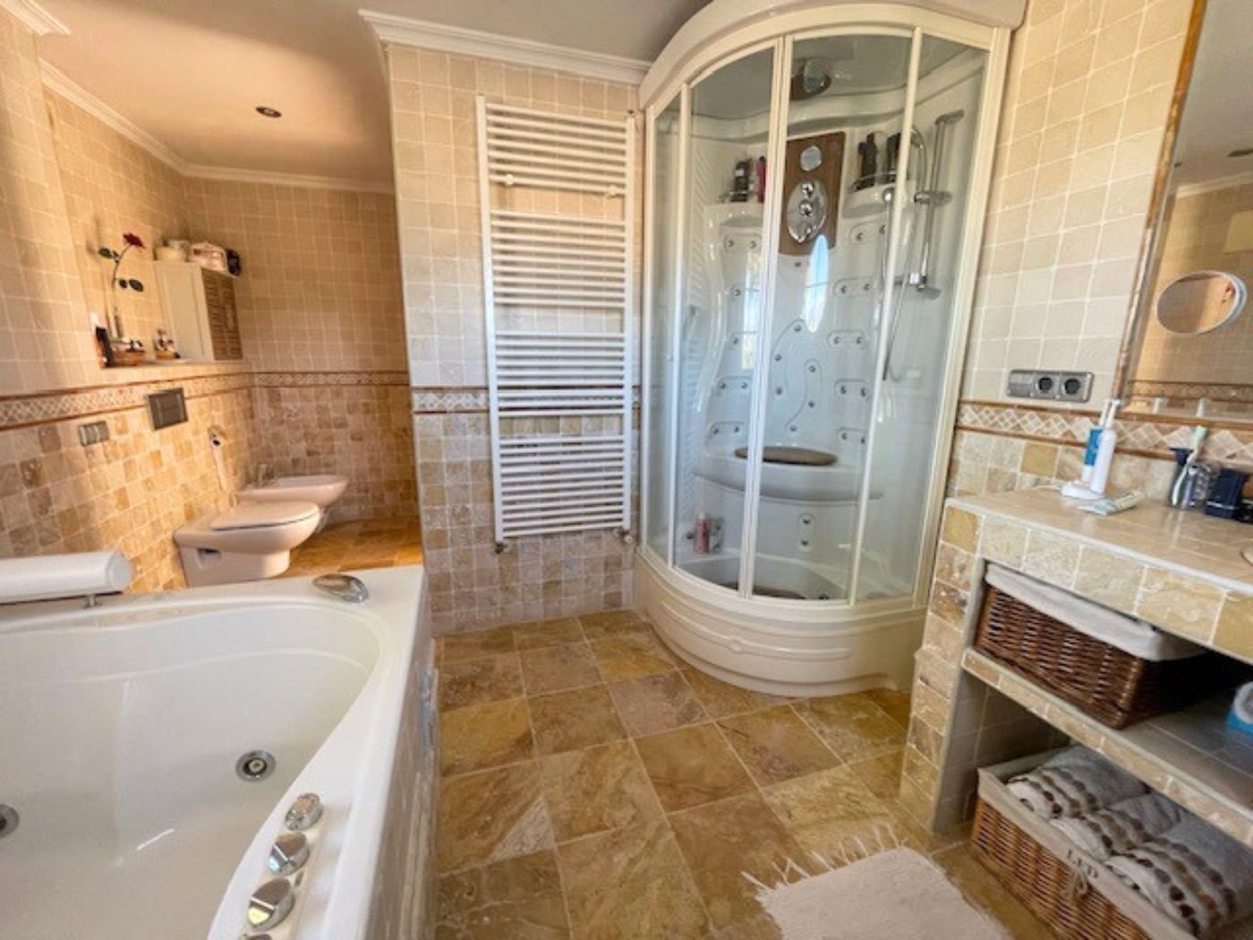 Badkamer met een hoekhydromassage-douchecabine, een ligbad en natuurstenen tegels. Voorzien van een witte handdoekradiator en opbergmanden van vlechtwerk.