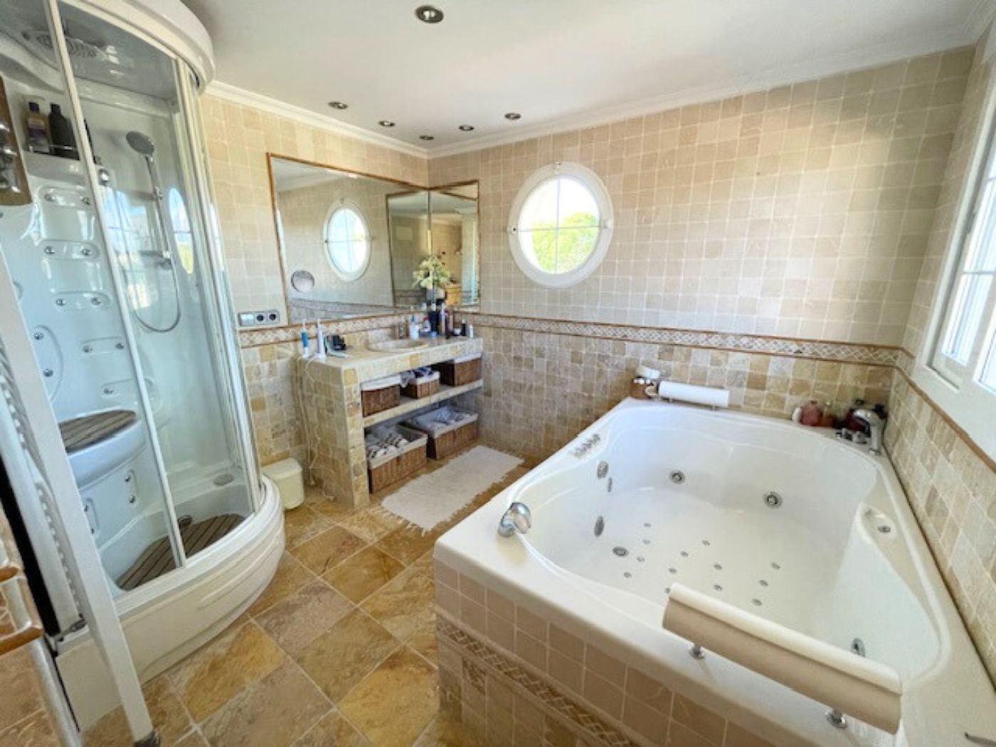 Badkamer met hydromassagebad, glazen douchecabine en betegelde wanden voorzien van een rond venster en inbouwspots.