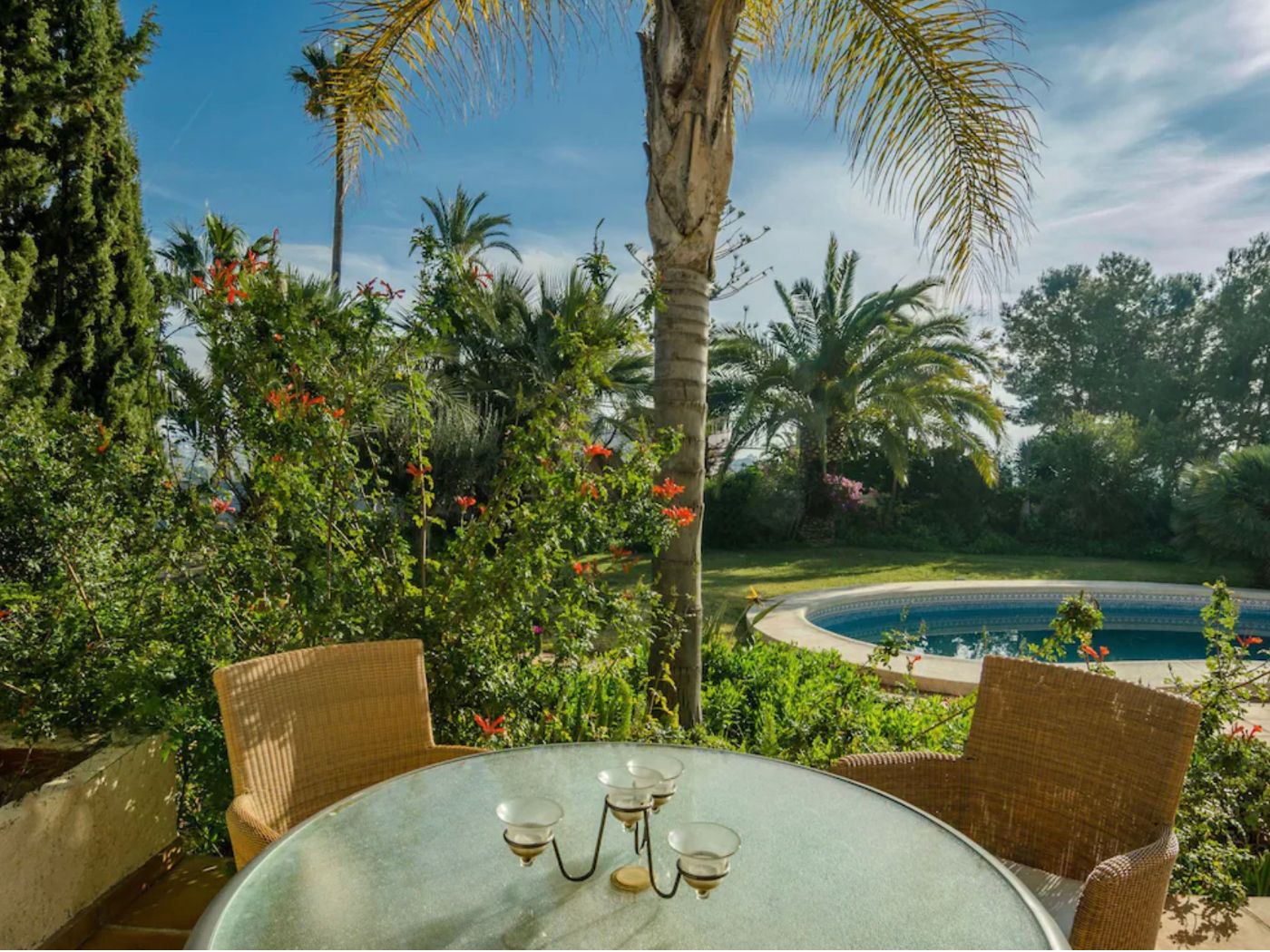 Buitenterras met een glazen tafel en rieten stoelen, uitkijkend op een rond zwembad en weelderige mediterrane tuinen met volwassen palmbomen.