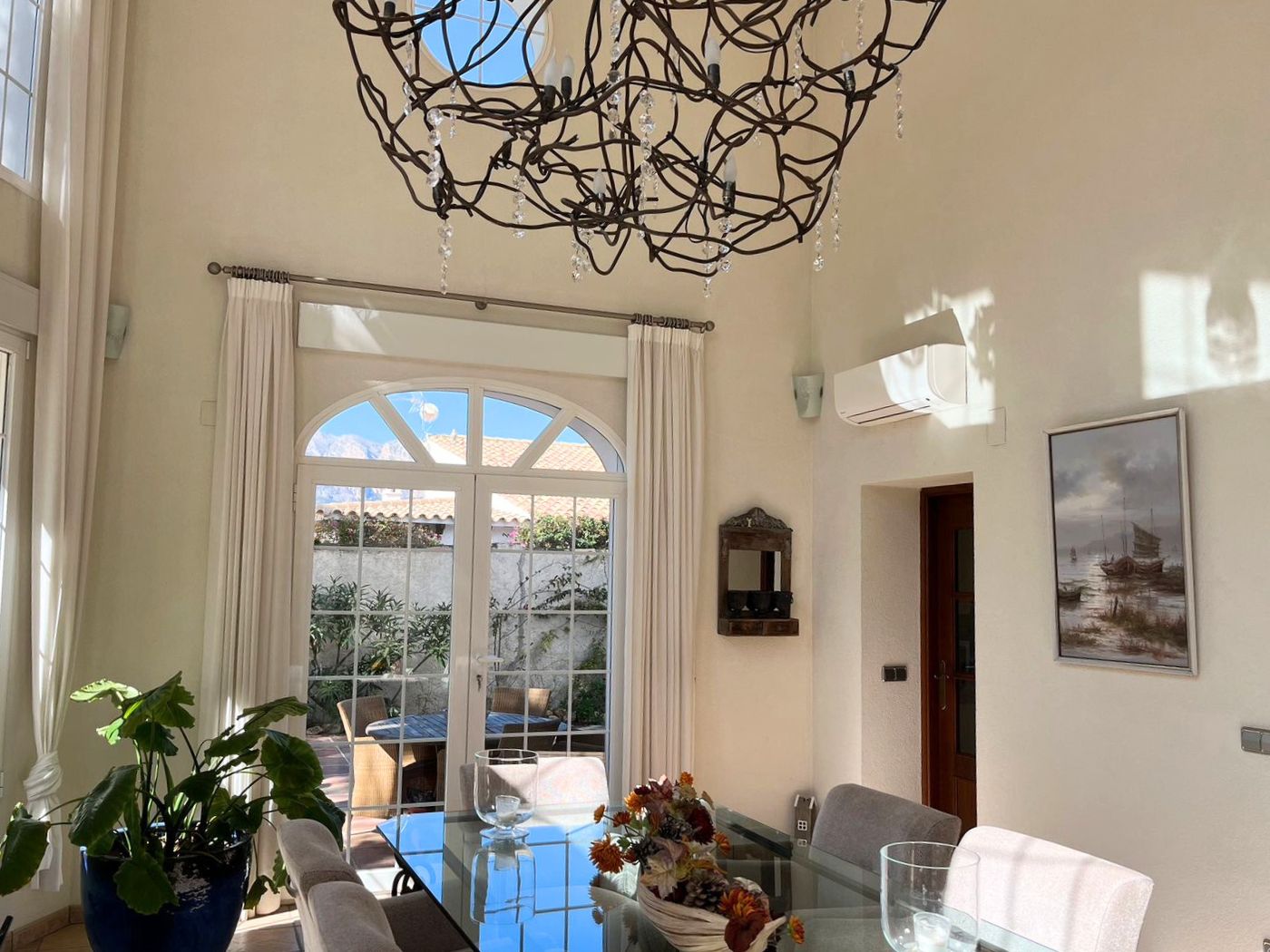 Eetkamer met hoog plafond, glazen tafel en een smeedijzeren kroonluchter. De boogdeuren geven toegang tot een zonnig terras met tuinmeubilair.