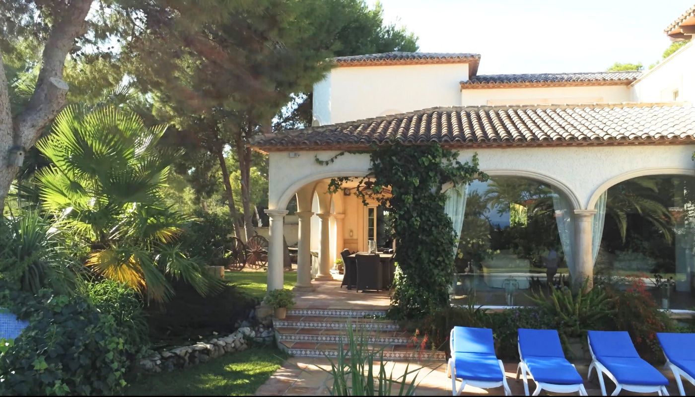 Exterieur van een mediterrane villa met een boogvormige veranda, stenen zuilen, een pannendak en een terras met blauwe ligbedden.