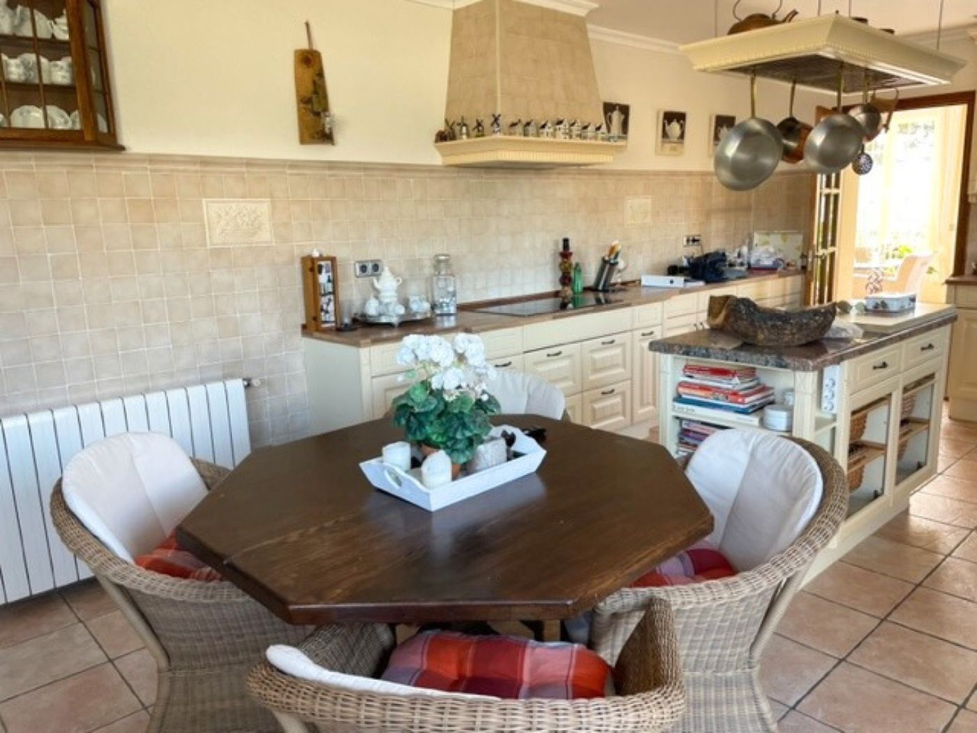 Keuken met een achthoekige houten tafel, rotan stoelen en een kookeiland met marmeren blad, afgewerkt met klassieke crèmekleurige kasten en tegelvloer.