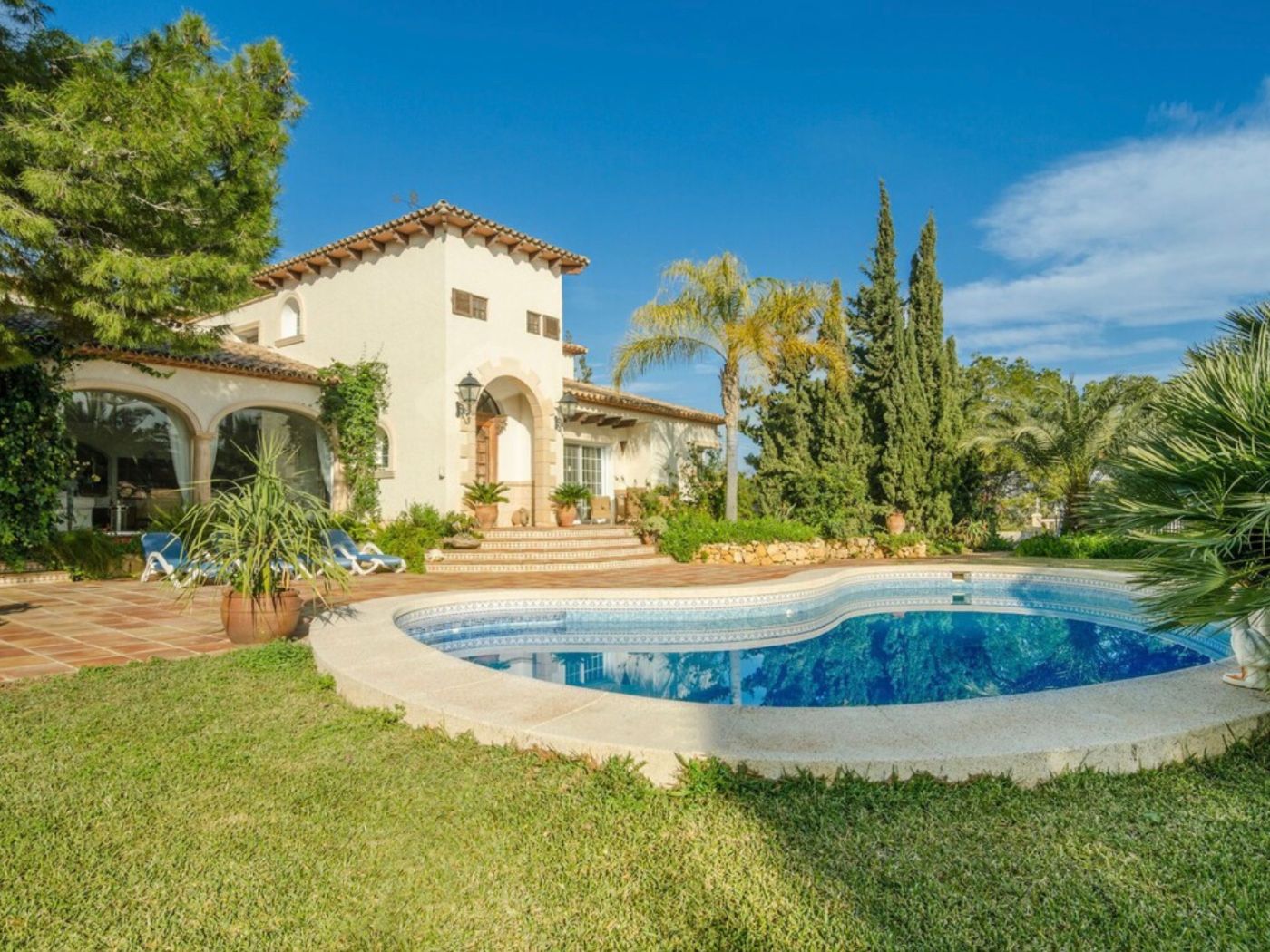 Mediterrane villa met een niervormig zwembad, terracotta terras en witte gevel met traditionele bogen, omgeven door een volgroeide tuin met palmen.