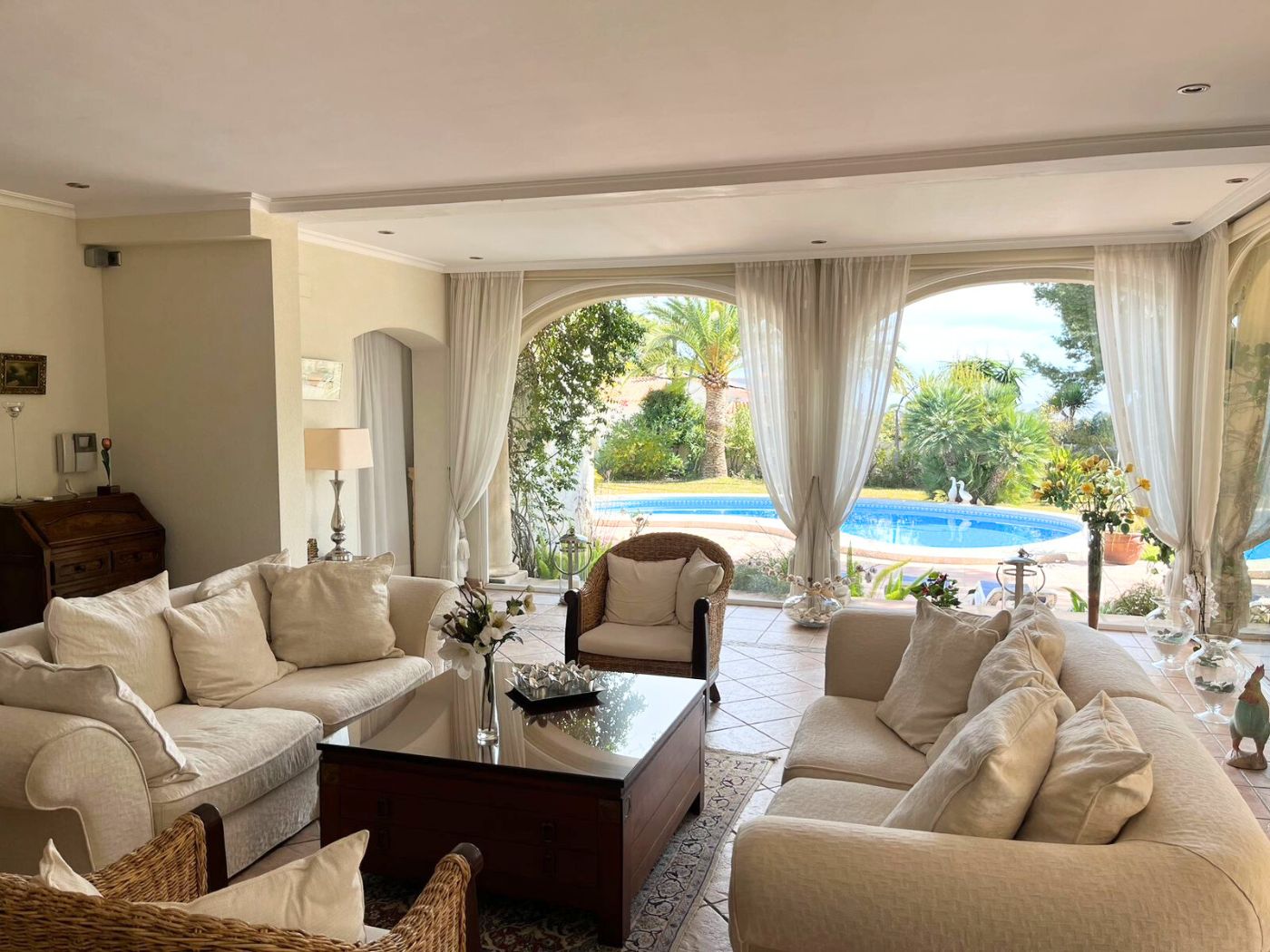 Ruime woonkamer met crèmekleurige banken en rotan fauteuils, voorzien van grote boogvensters die uitkijken op het zwembad en de mediterrane tuin.