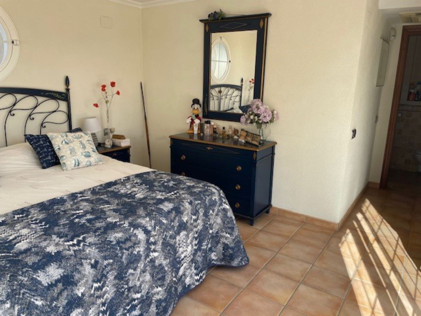 Slaapkamer met een smeedijzeren bedframe, een marineblauwe ladekast met bijbehorende spiegel en een traditionele terracotta tegelvloer met natuurlijk licht.