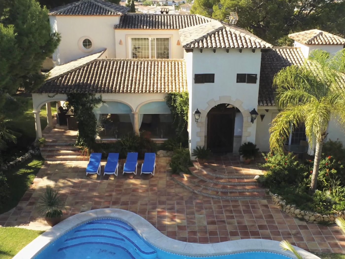 Verhoogd zicht op een mediterrane villa met een gebogen zwembad, terracotta terras met vier ligstoelen en een witte gevel met stenen boogingangen.