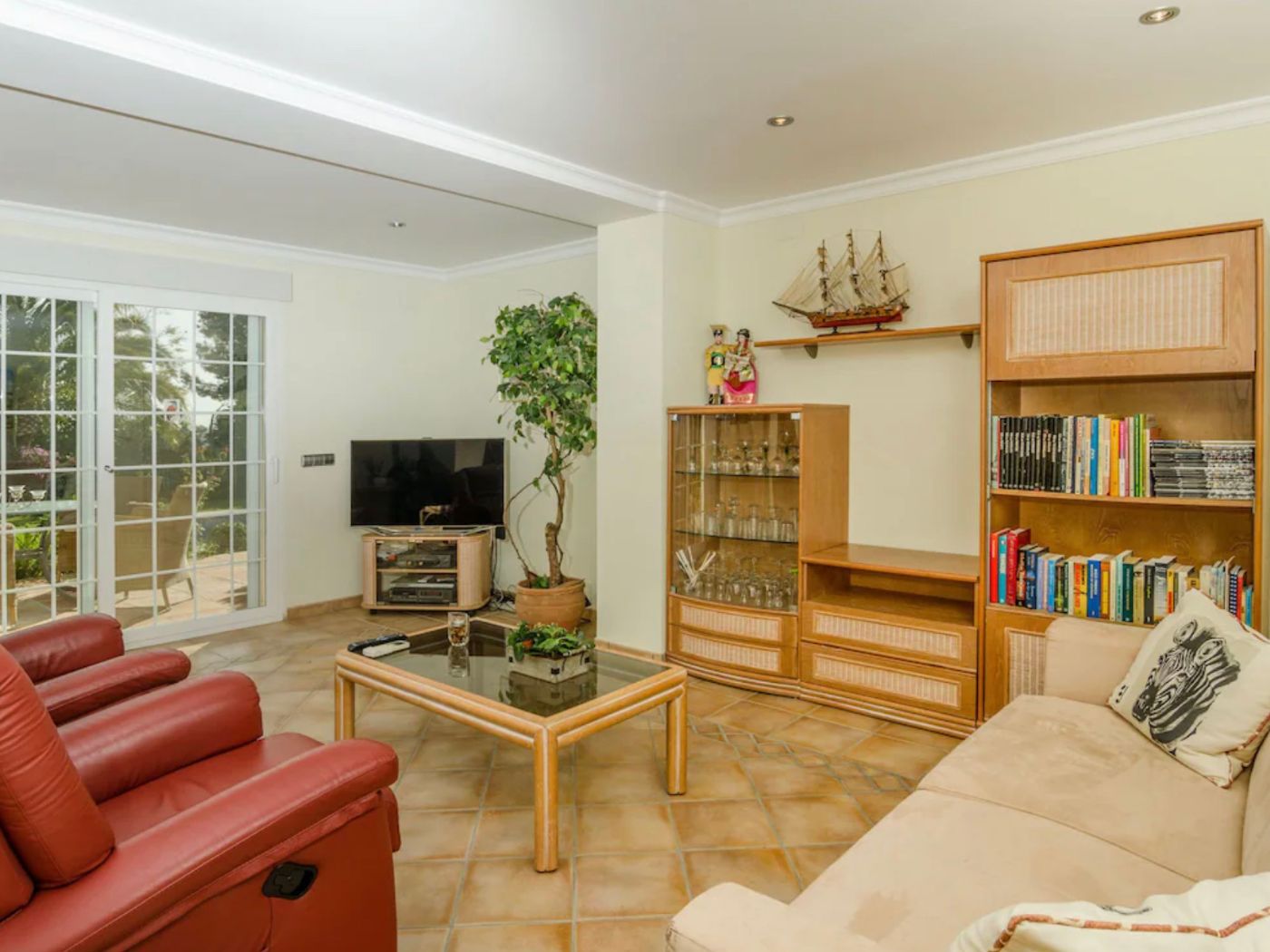 Woonkamer met terracotta vloertegels, rode lederen fauteuils en houten kastenwand. Grote glazen schuifdeuren bieden toegang tot het terras.