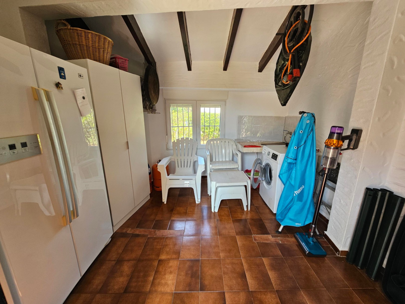 Bijkeuken met een terracotta tegelvloer, donkere houten plafondbalken, een wasmachine en witte inbouwkasten voor extra opslag.