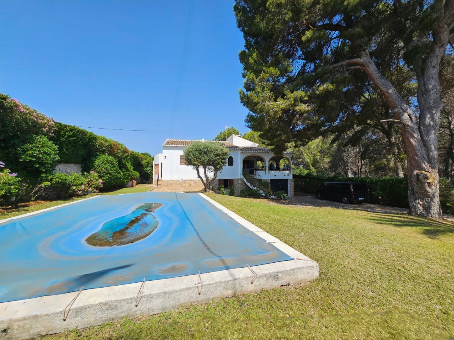 Buitenaanzicht van een witte villa met een boogterras en een groot gazon met een afgedekt zwembad, omgeven door volwassen pijnbomen en mediterrane beplanting.