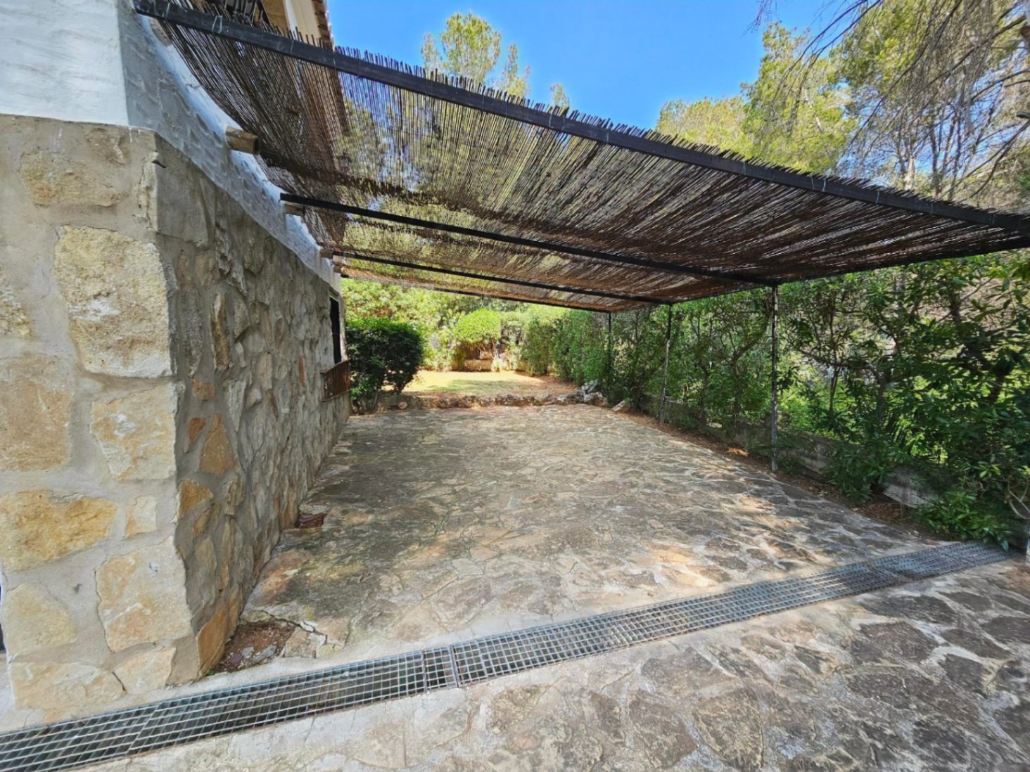 Buitenterras met natuurstenen bestrating en een rustieke rieten pergola, grenzend aan een stenen gevel en omgeven door mediterrane begroeiing.