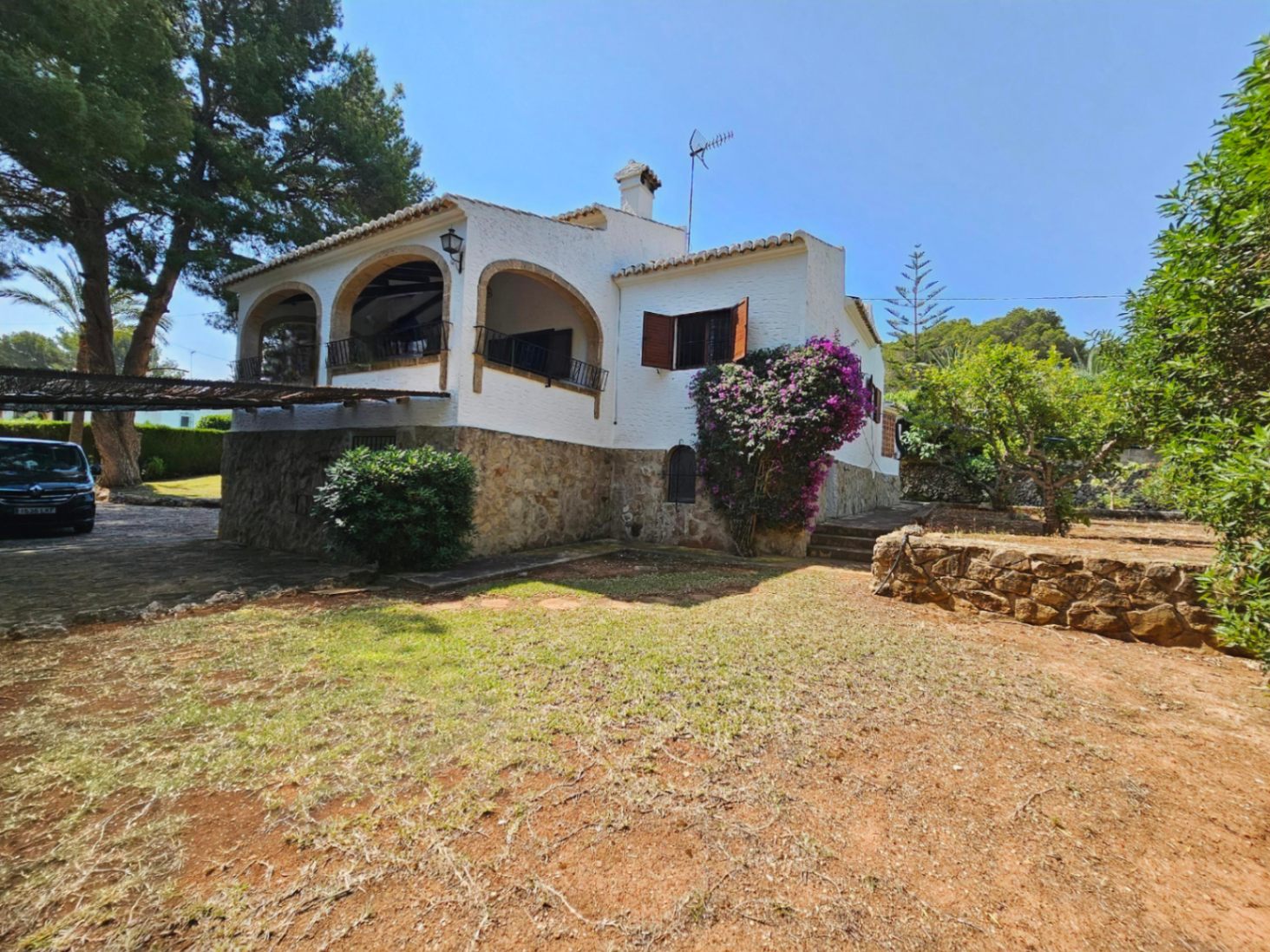 Exterieur van een mediterrane villa met natuurstenen plint, witgepleisterde muren en een overdekt terras met bogen. Tuin met volwassen bomen en bougainville.