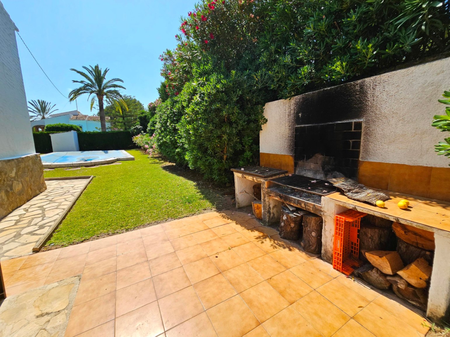 Gemetselde buitenbarbecue met betegelde werkbladen en houtopslag, gelegen naast een gazon en een zwembad omgeven door mediterrane beplanting.