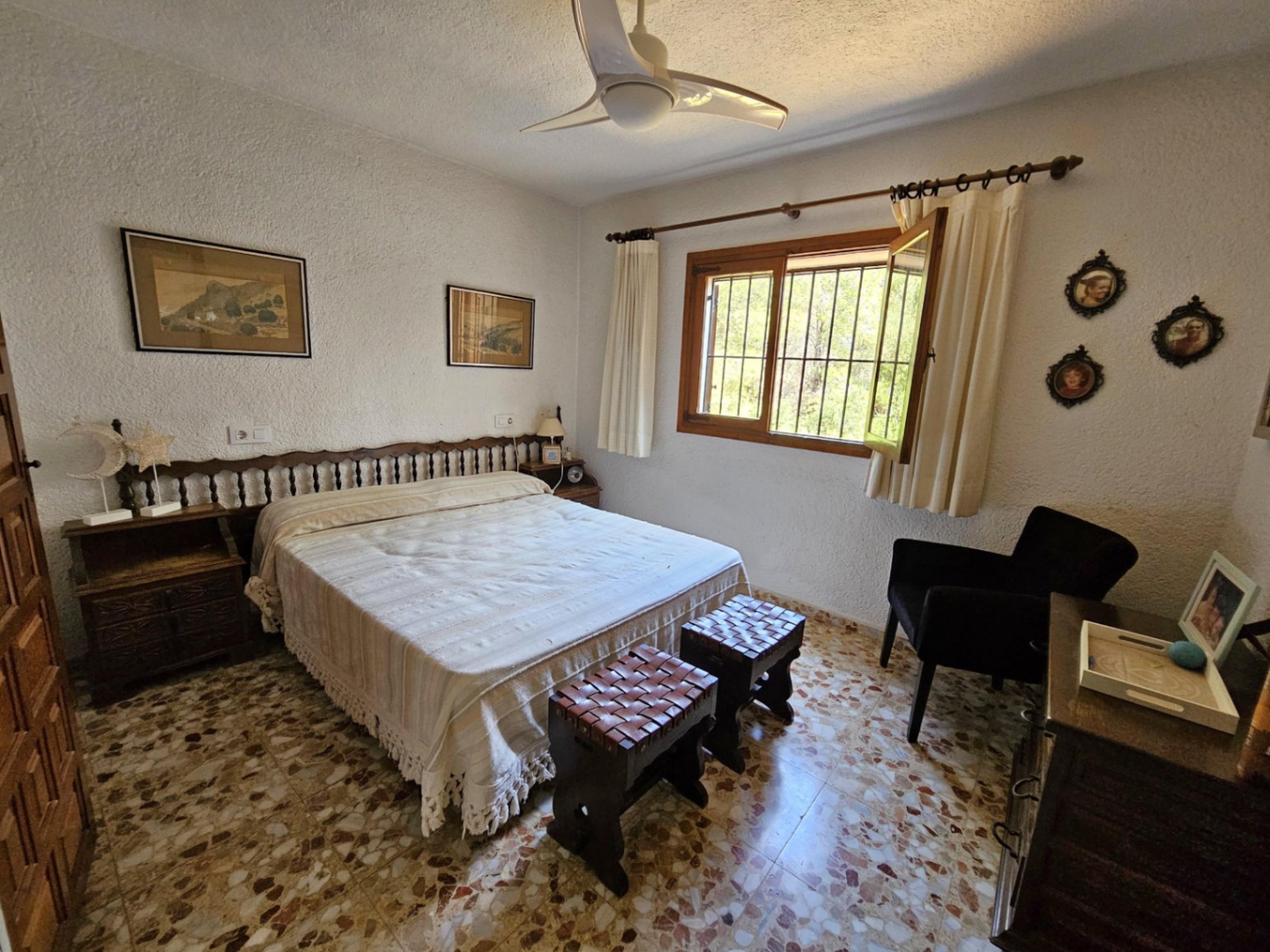 Slaapkamer met traditionele terrazzovloer, houten spijlen hoofdbord en krukjes met gevlochten leer. Voorzien van een witte plafondventilator en houten raamkozijn.