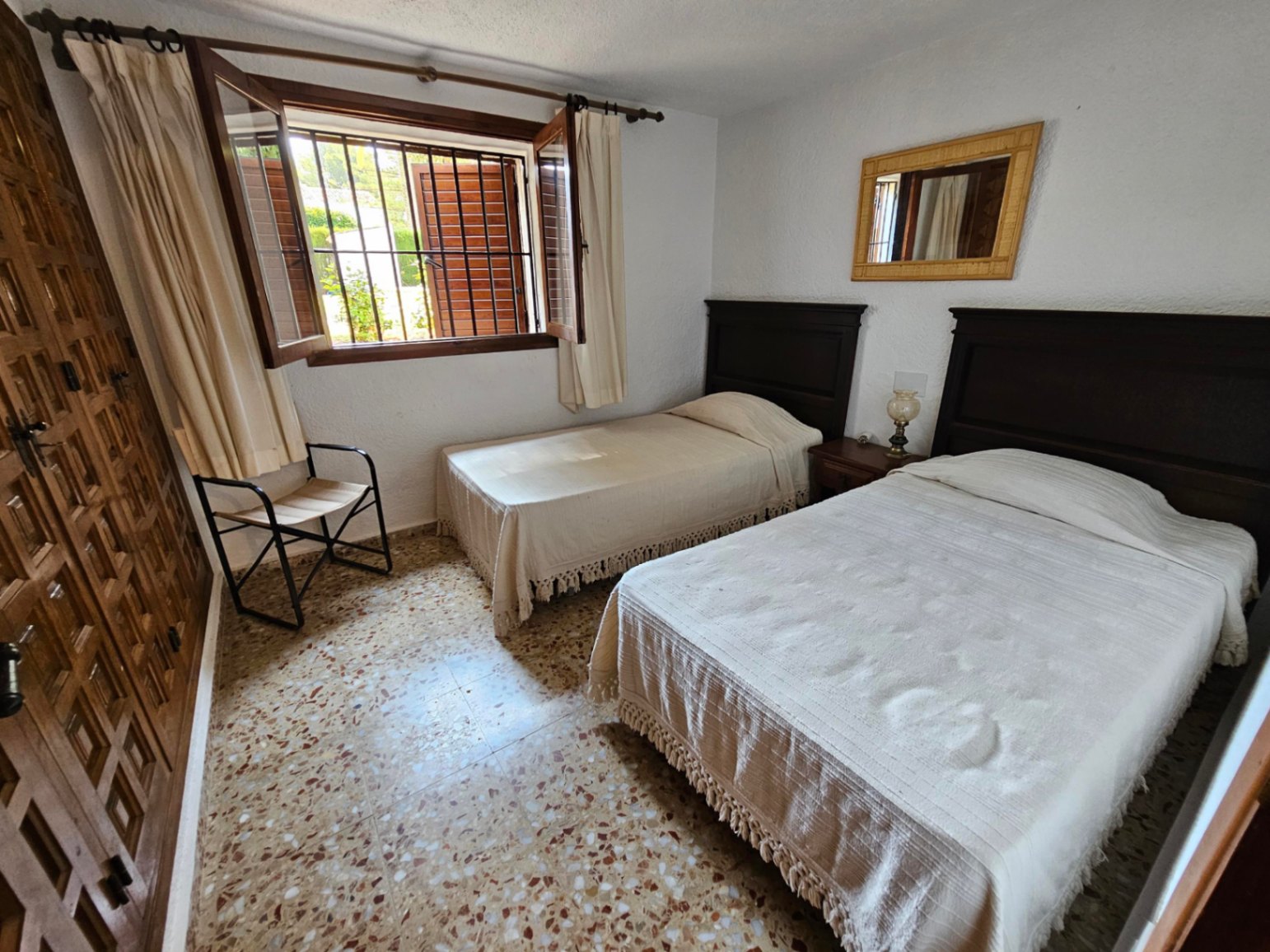 Slaapkamer met twee eenpersoonsbedden, donkerhouten hoofdeinden, een traditionele terrazzovloer en een houten raam met luiken en beveiligingsstangen.