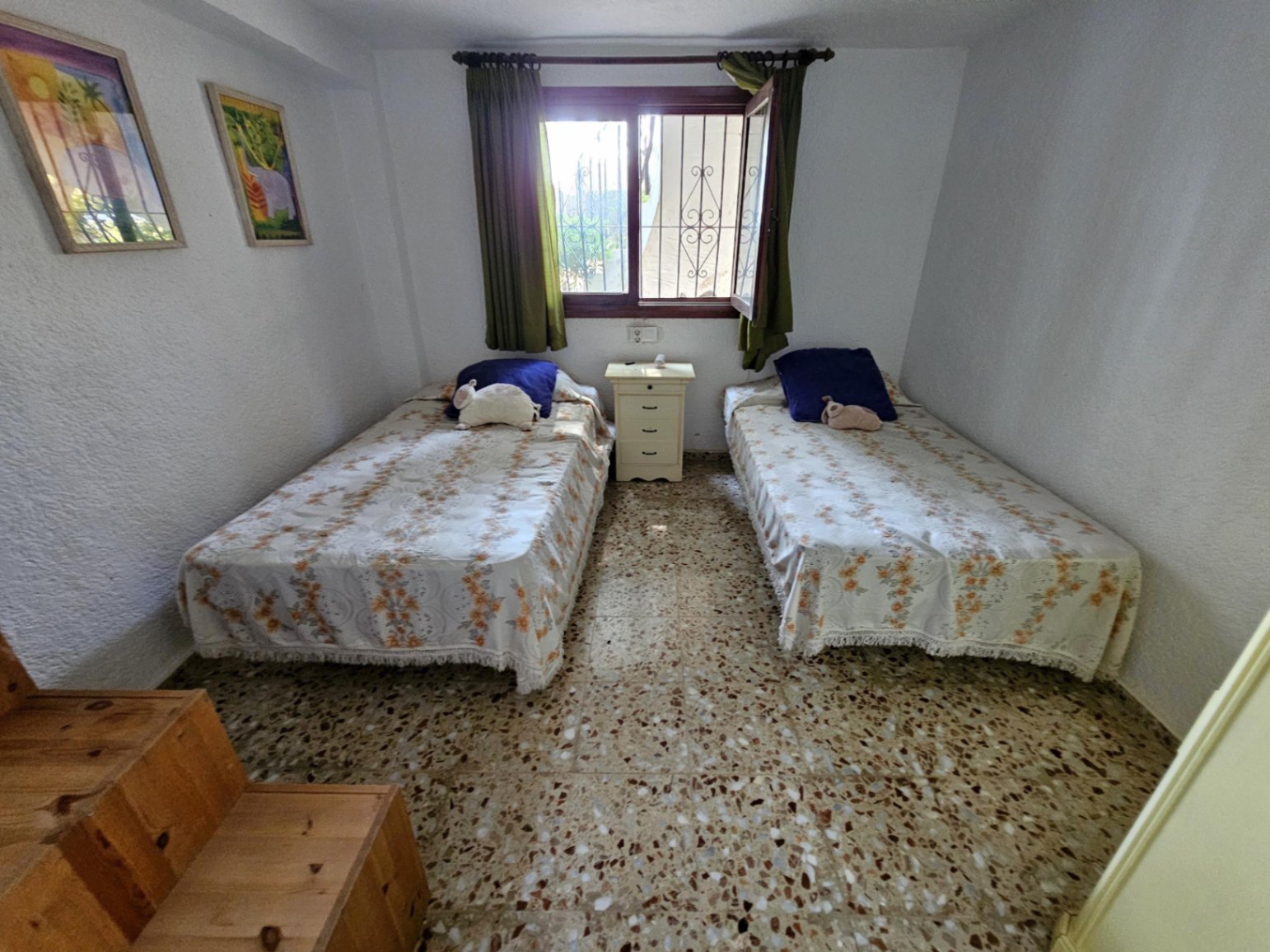 Slaapkamer met twee eenpersoonsbedden, een terrazzovloer en een centraal raam met tralies en groene gordijnen. Tussen de bedden staat een wit nachtkastje.