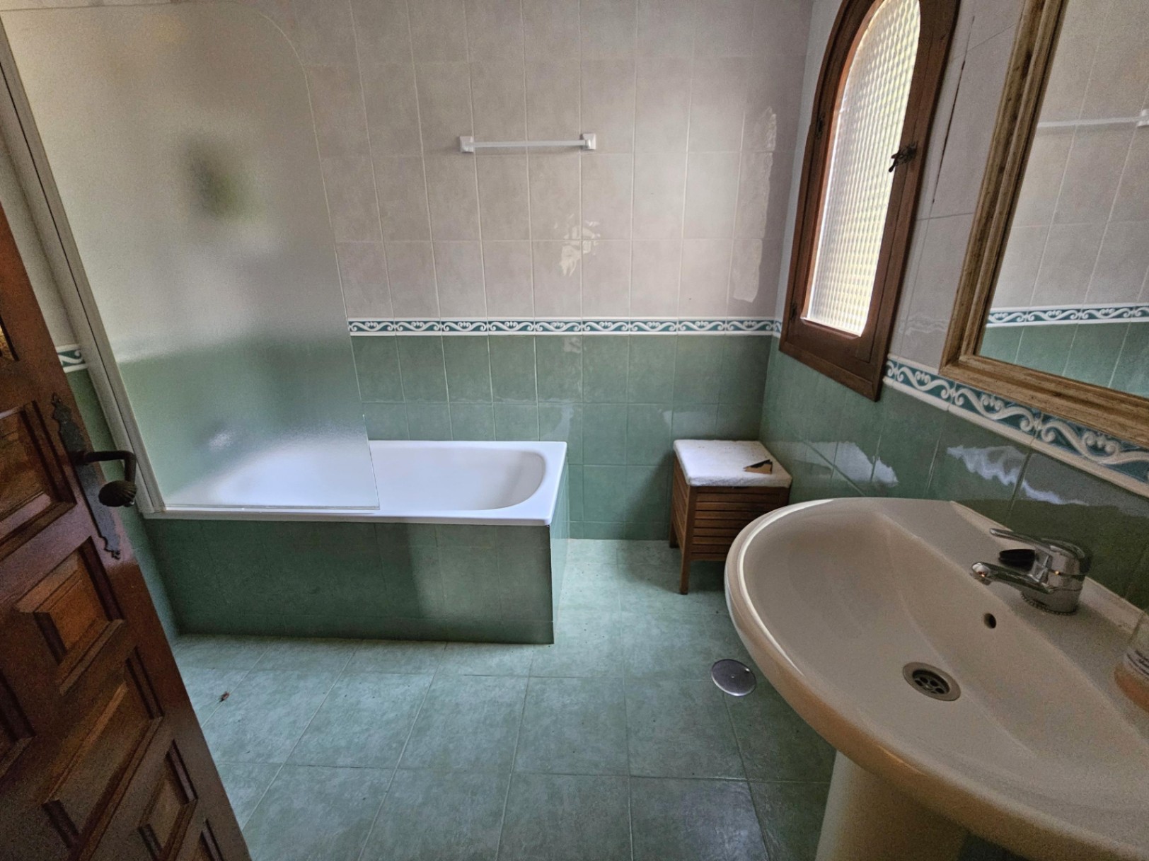 Traditionele badkamer met groen-witte betegeling, een ingebouwd ligbad met matglazen wand en een klassieke wastafel op zuil bij een houten boograam.