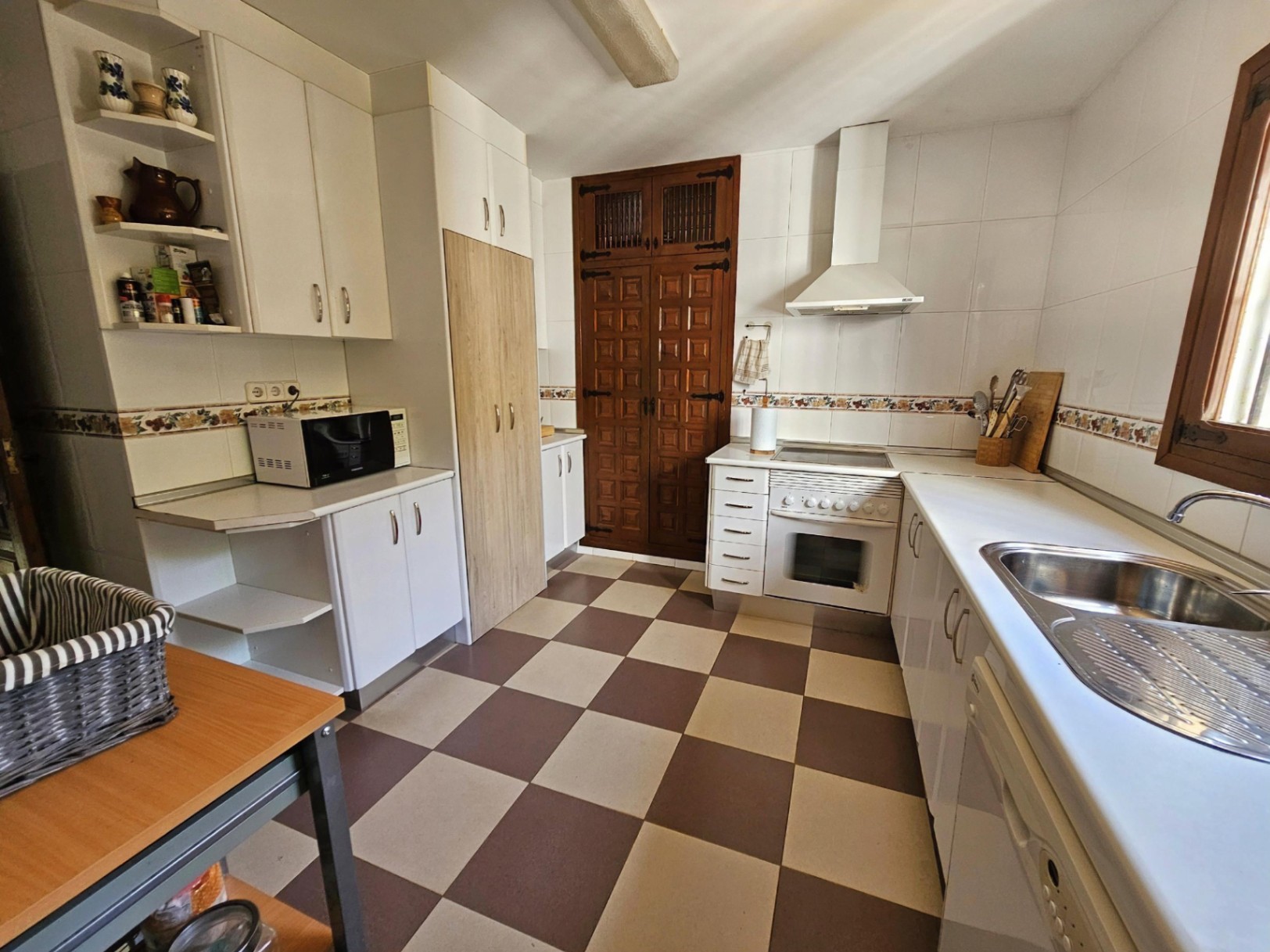 Traditionele keuken met een geblokte tegelvloer, witte kasten en een fraai bewerkte houten deur. Voorzien van een rvs spoelbak en witte apparatuur.