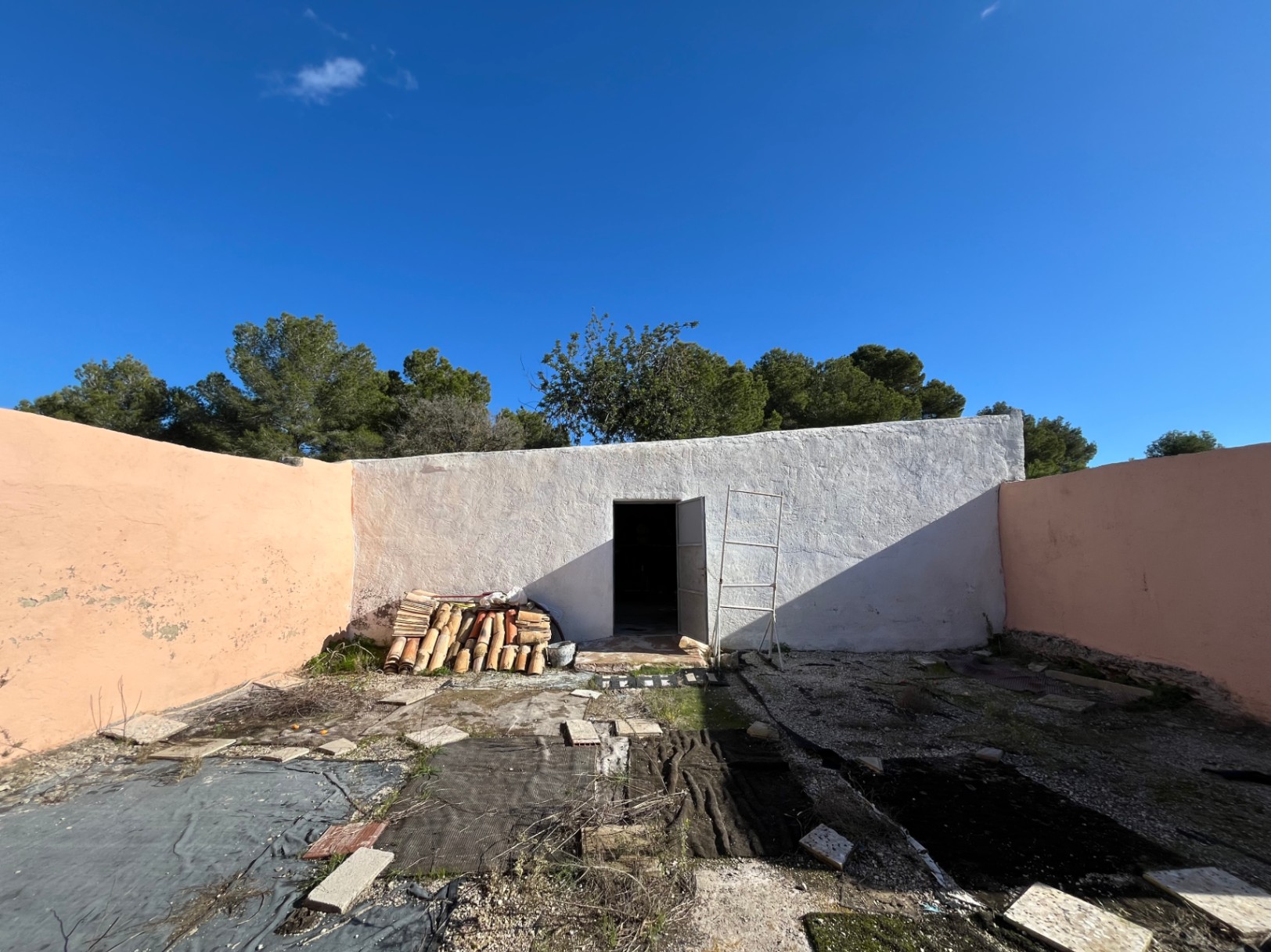 Besloten binnenplaats met witgekalkt bijgebouw, stapels traditionele terracotta dakpannen en okerkleurige muren onder een strakblauwe lucht.