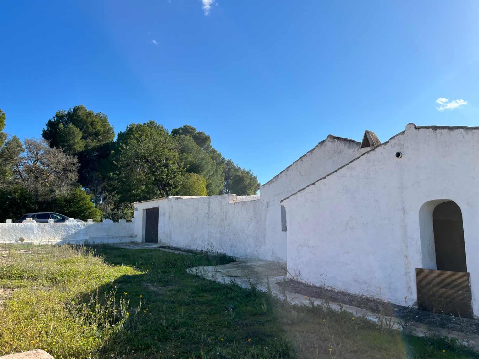 Buitenaanzicht van een traditionele witgekalkte finca in Jávea, met een boogingang, authentieke muren en omringende pijnbomen op een rustiek perceel.