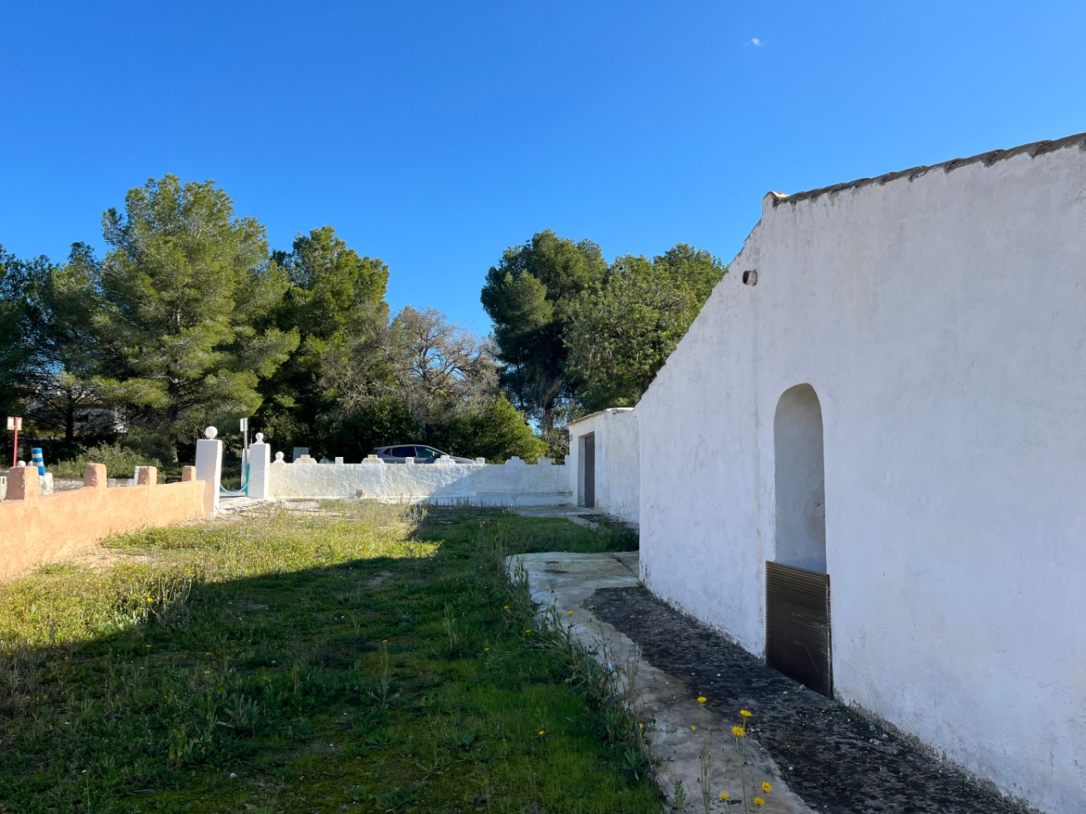 Buitenzijde van een traditionele witte finca in Jávea met een boognis en een natuurlijk perceel, omzoomd door pijnbomen en een witte tuinmuur.