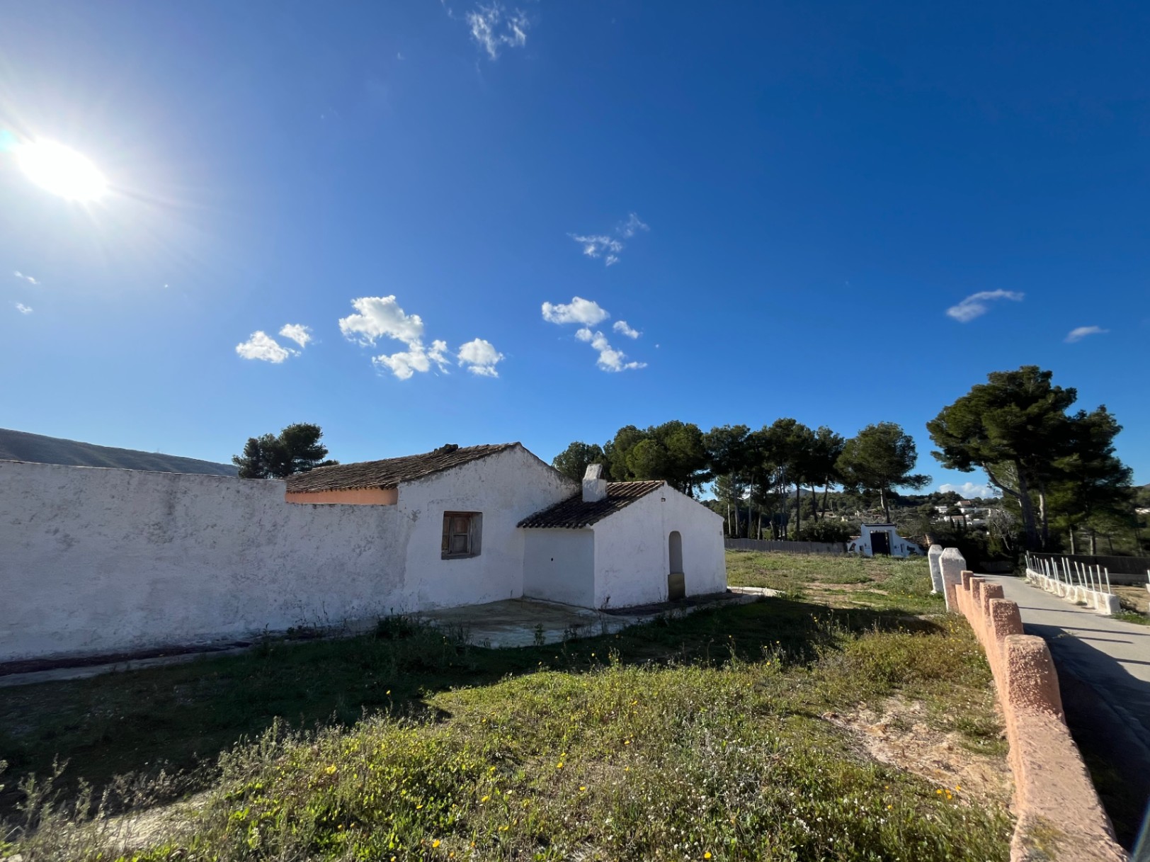 Exterieur van een traditionele witgekalkte finca met pannendak, gelegen op een natuurlijk perceel met pijnbomen onder een heldere blauwe lucht.