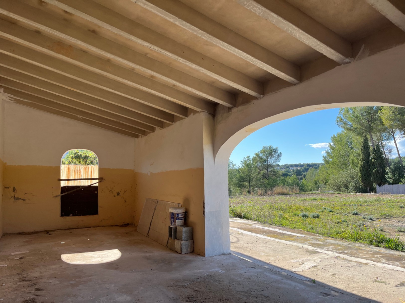 Te renoveren veranda met balkenplafond en een grote boog die uitkijkt over de omliggende velden en mediterrane vegetatie.