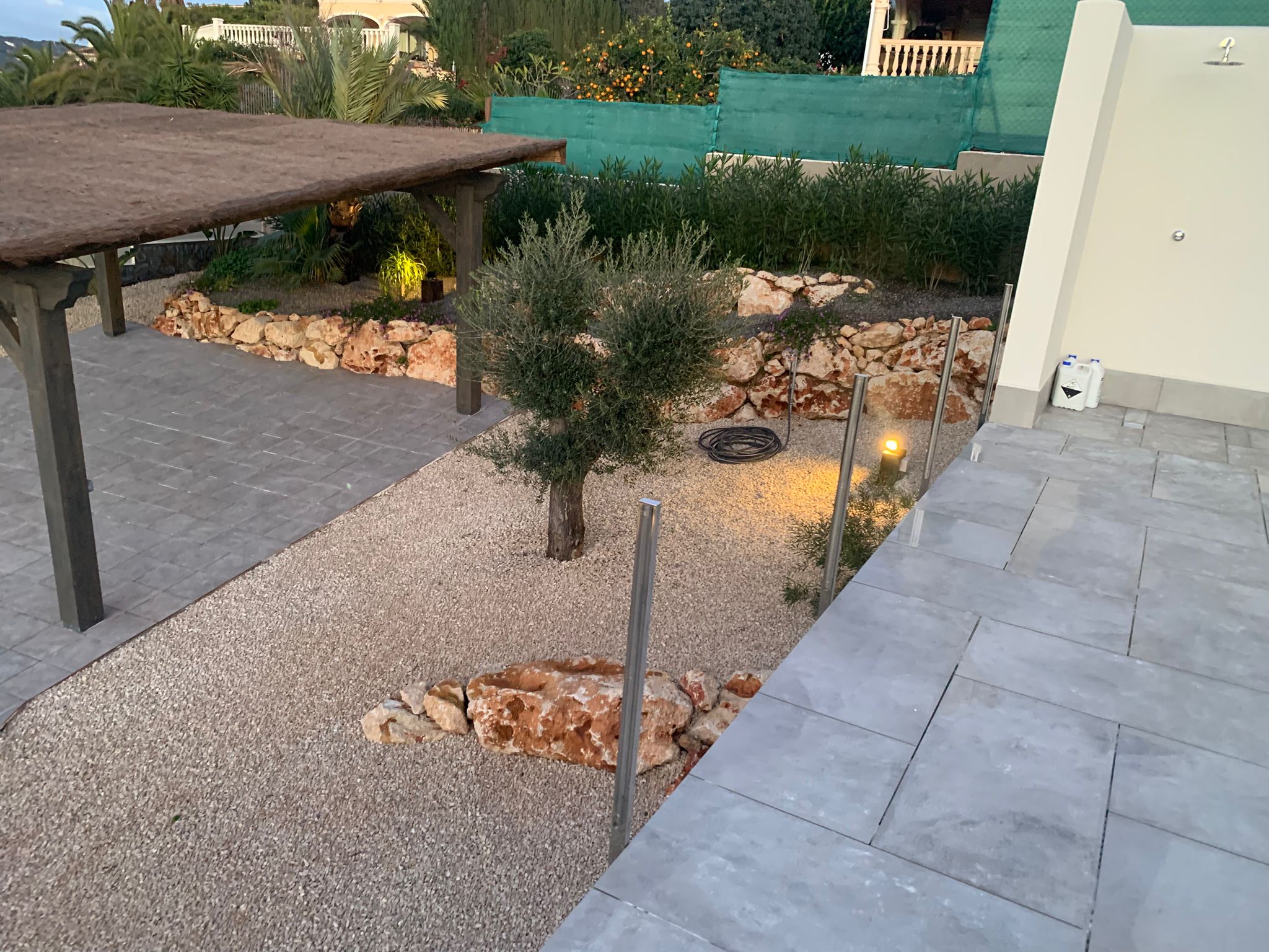 Mediterrane tuin met grind, een olijfboom, natuurstenen borders, een rieten pergola en grijs betegeld terras met buitendouche.
