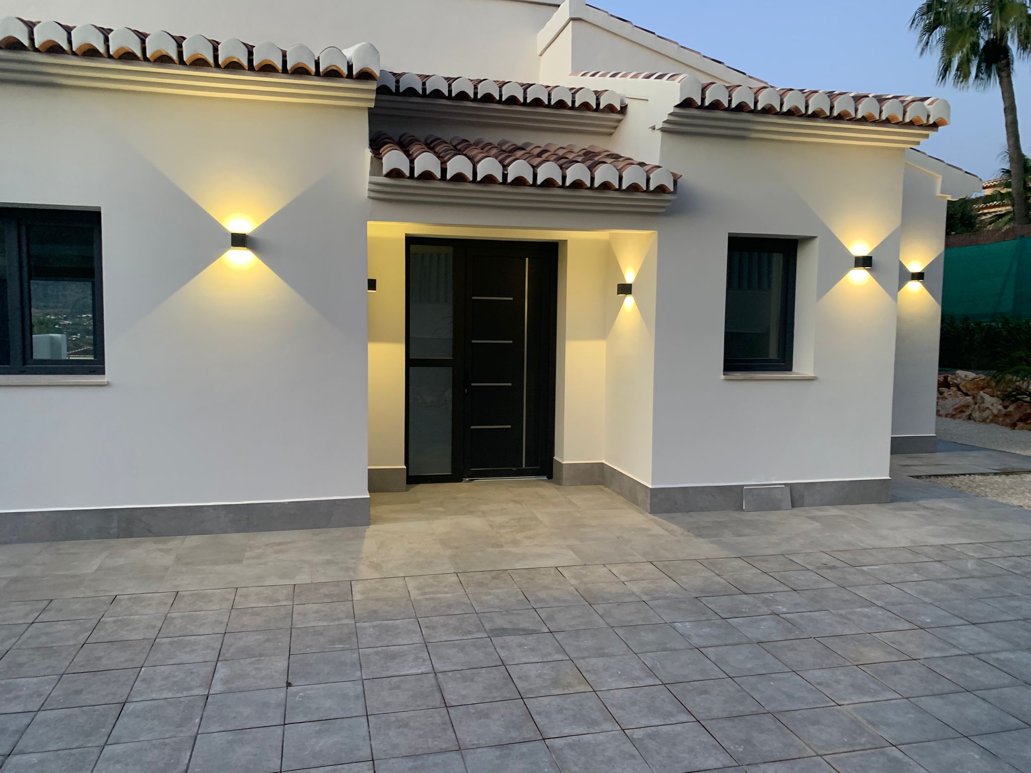 Moderne villa-entree met een zwarte deur, witte gevel en grijs betegeld terras. Symmetrische wandarmaturen zorgen voor warme verlichting onder het pannendak.