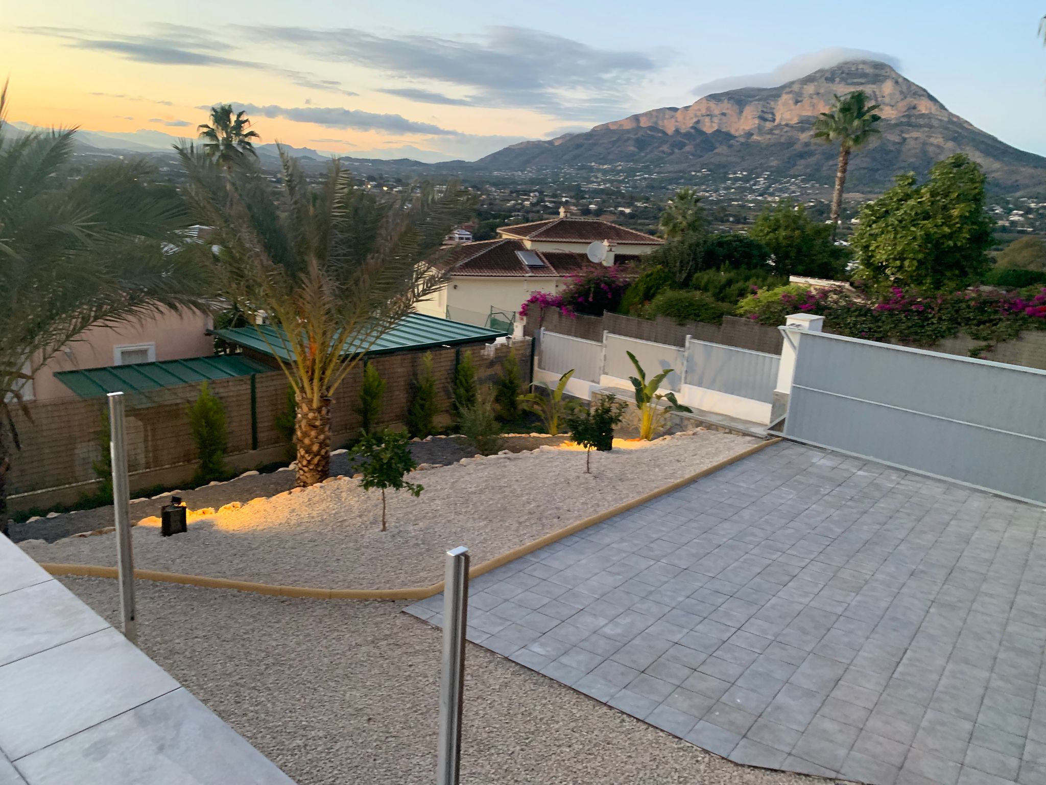 Verhoogd uitzicht op een aangelegde tuin met grind, palmbomen en een geplaveide oprit, met panoramisch uitzicht op de bergen bij zonsondergang.