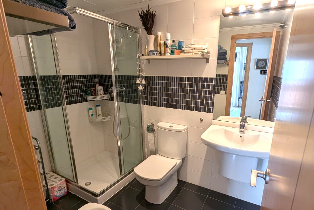 Badkamer met glazen douchecabine, witte wandtegels met een donkere mozaïekrand en een antracietgrijze tegelvloer.