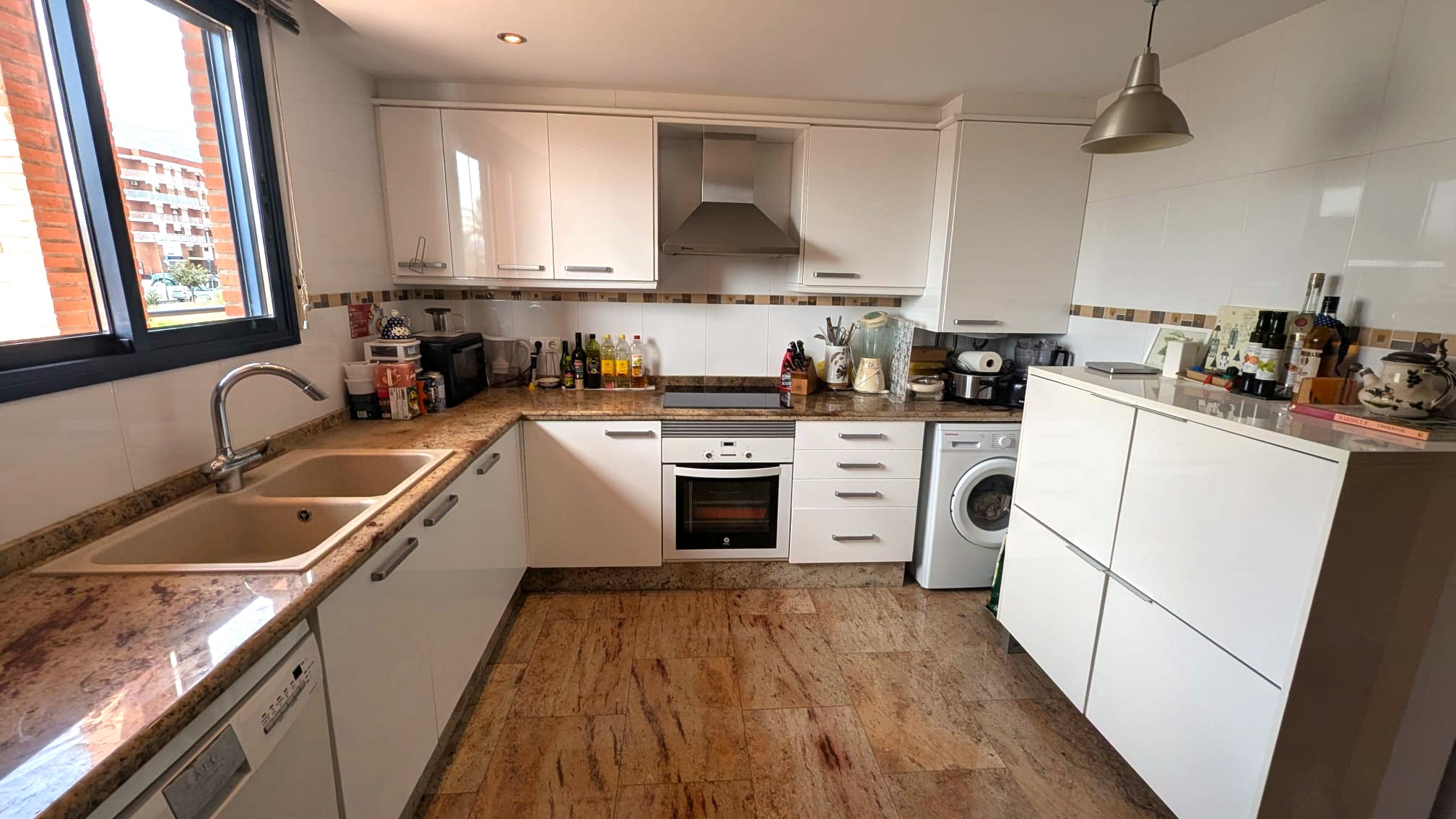 Functionele keuken met witte kasten, granieten werkbladen en een natuurstenen vloer. Voorzien van inbouwapparatuur en een raam met donker kozijn.