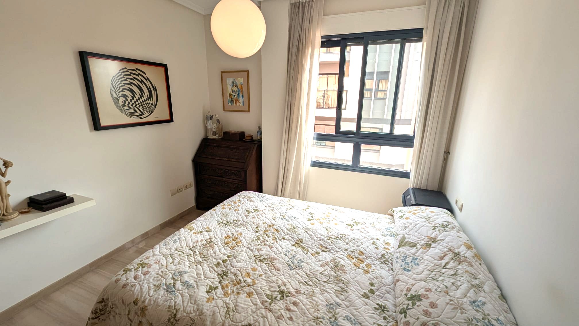 Lichte slaapkamer met een antiek houten bureau, een groot raam met lichte gordijnen en een minimalistische bolvormige hanglamp boven het bed.