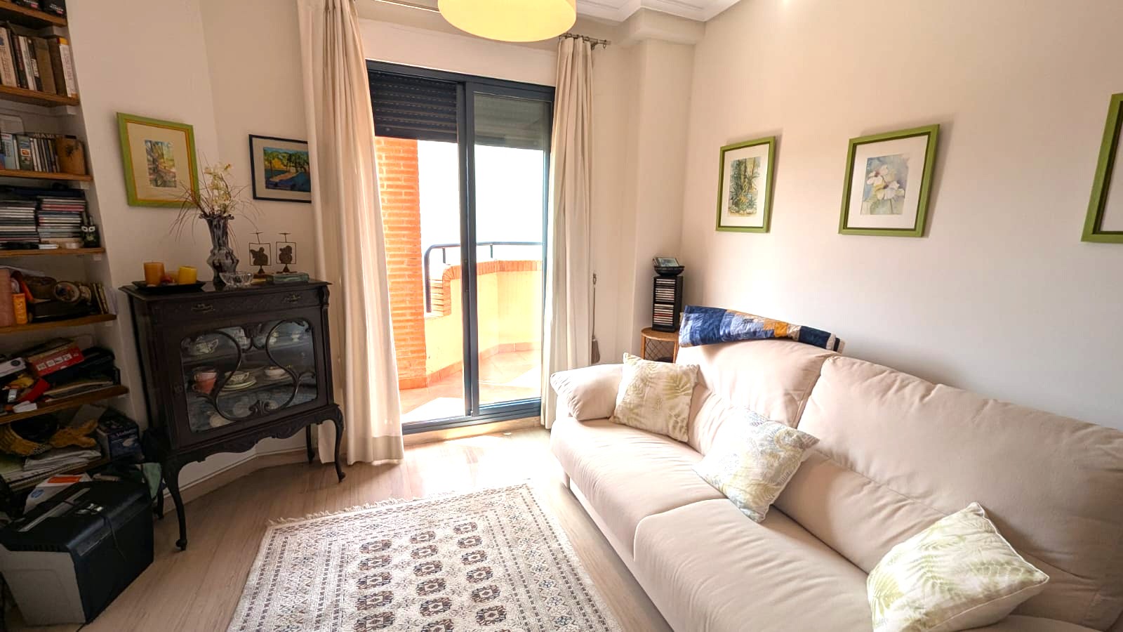 Lichte woonkamer met beige bank, klassieke donkerhouten vitrinekast en schuifpui die toegang biedt tot een balkon met bakstenen muur.