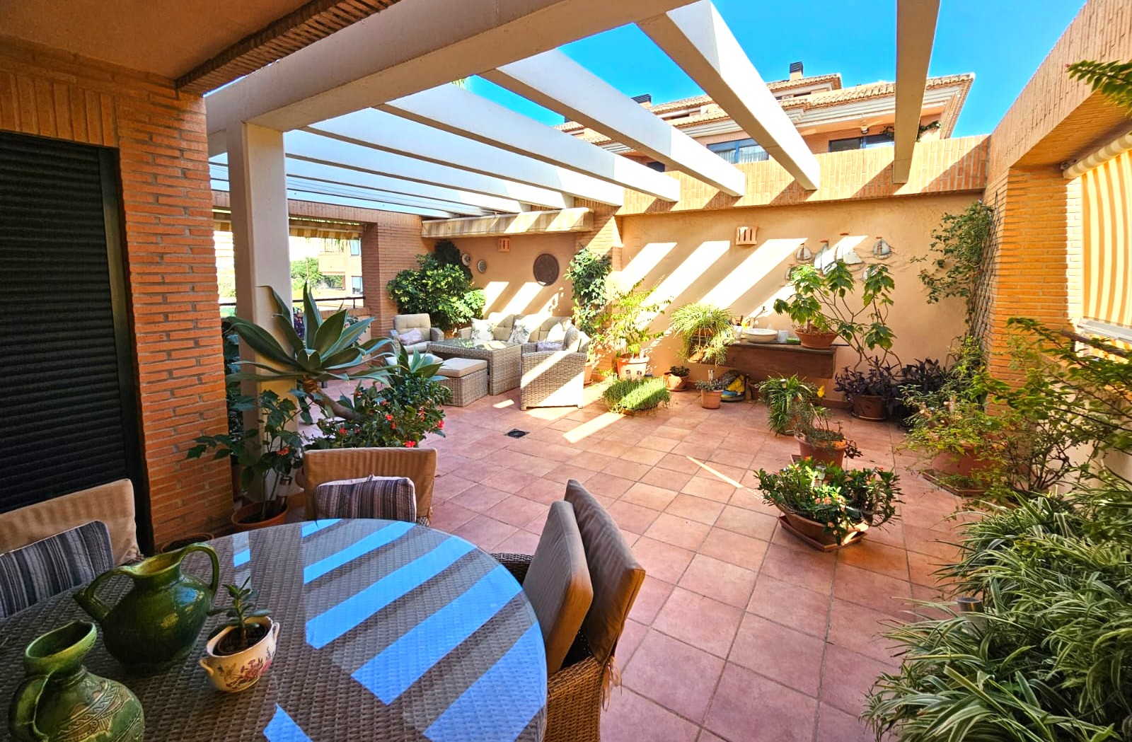 Ruim mediterraan terras met terracotta tegels, een witte pergola en diverse zithoeken omgeven door weelderige plantenbakken en natuurlijke schaduw.