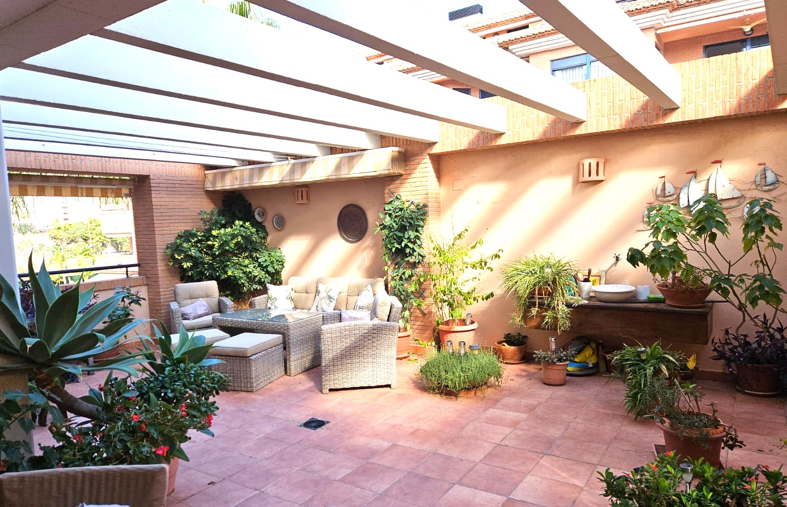 Ruim privéterras met terracotta tegels en witte pergolabalken. Voorzien van een wicker loungeset, bakstenen muren en diverse mediterrane potplanten.