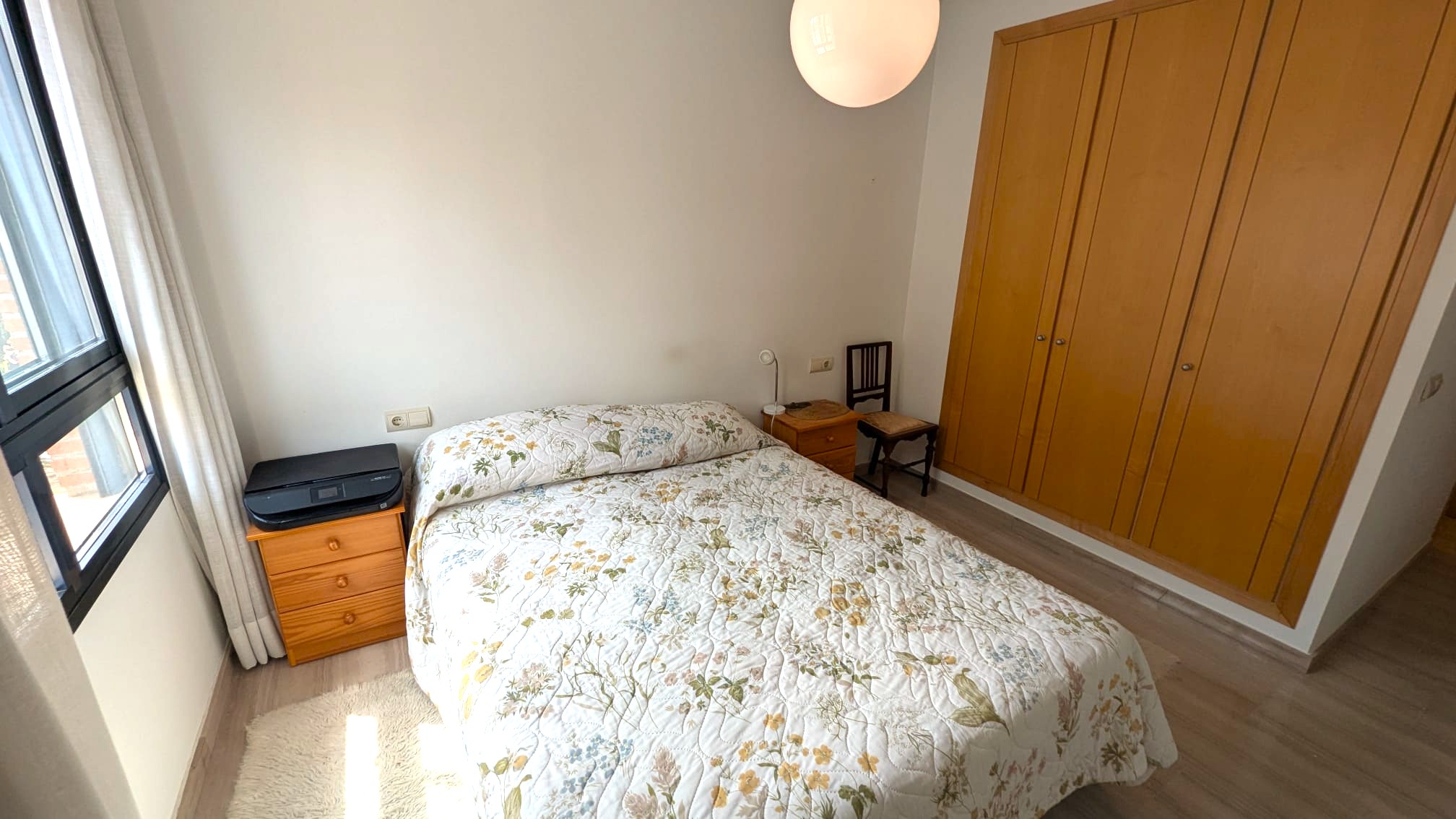 Slaapkamer met een ingebouwde vierdeurs houten kledingkast, tweepersoonsbed en bijpassende nachtkastjes. Voorzien van een lichte vloer en raam met zwart kozijn.