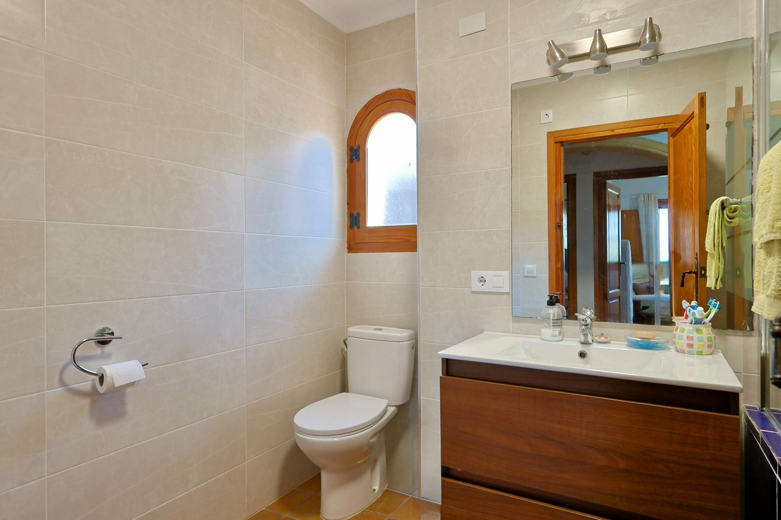 Badkamer met donkerhouten wastafelmeubel, wit wasbekken en een karakteristiek houten boogvenster tegen een achtergrond van beige wandtegels.