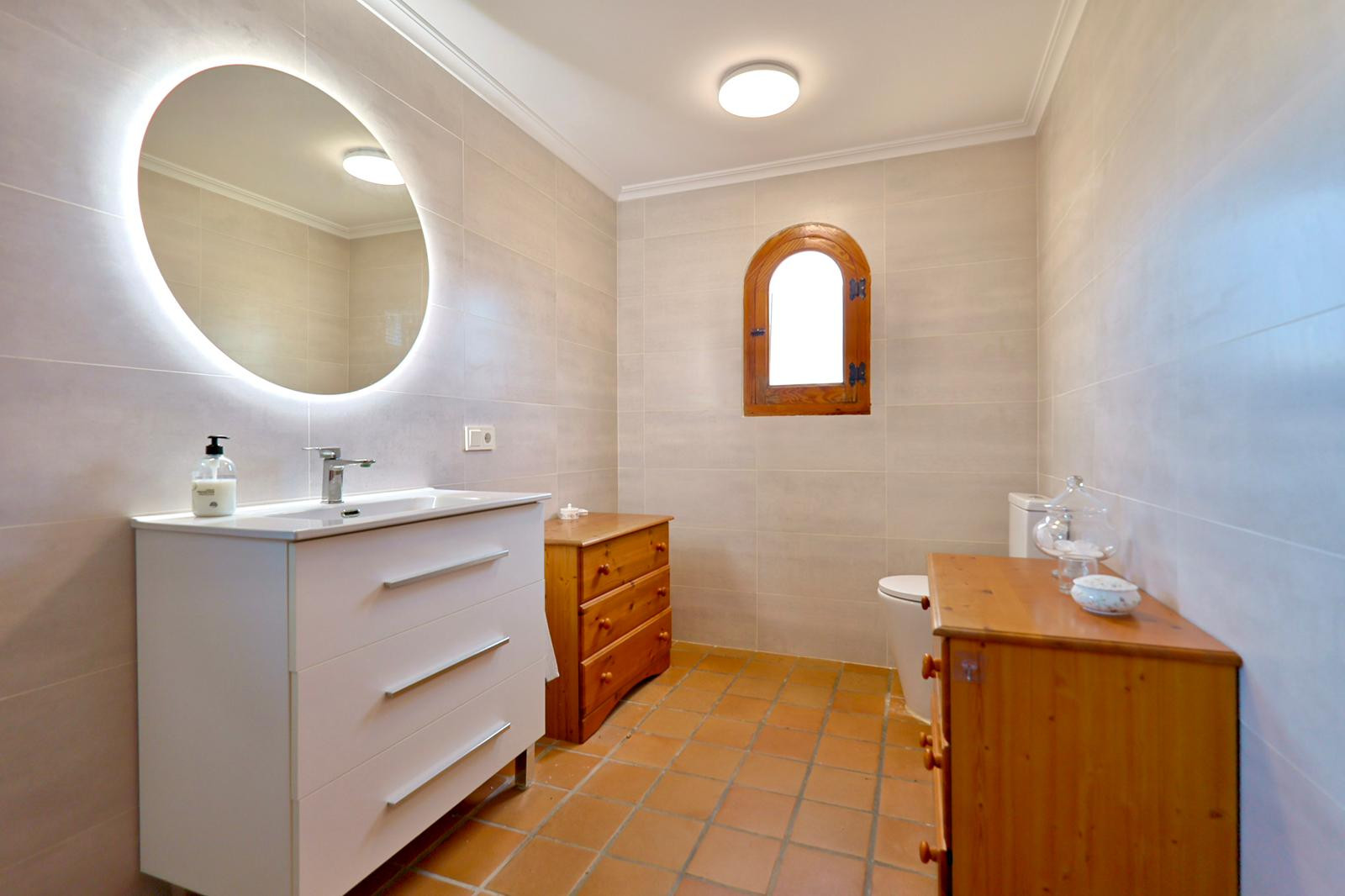 Badkamer met wit wastafelmeubel, ronde spiegel met ledverlichting en terracotta vloertegels. Voorzien van een houten boogvenster en grenen ladekasten.