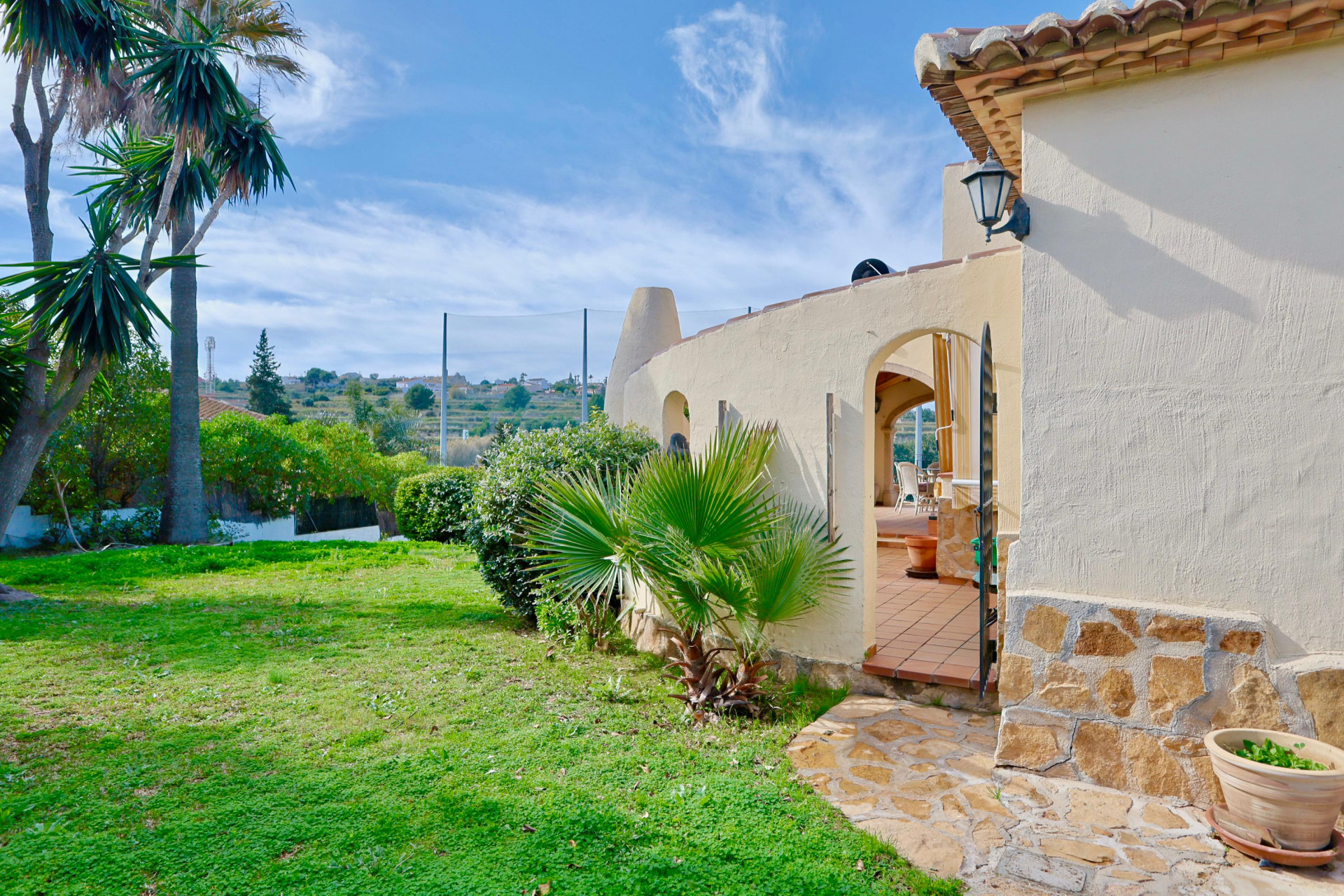 Buitengevel van de villa met natuurstenen plint, mediterrane tuin en een boogingang naar het terracotta terras met uitzicht op de golfbaanomgeving.