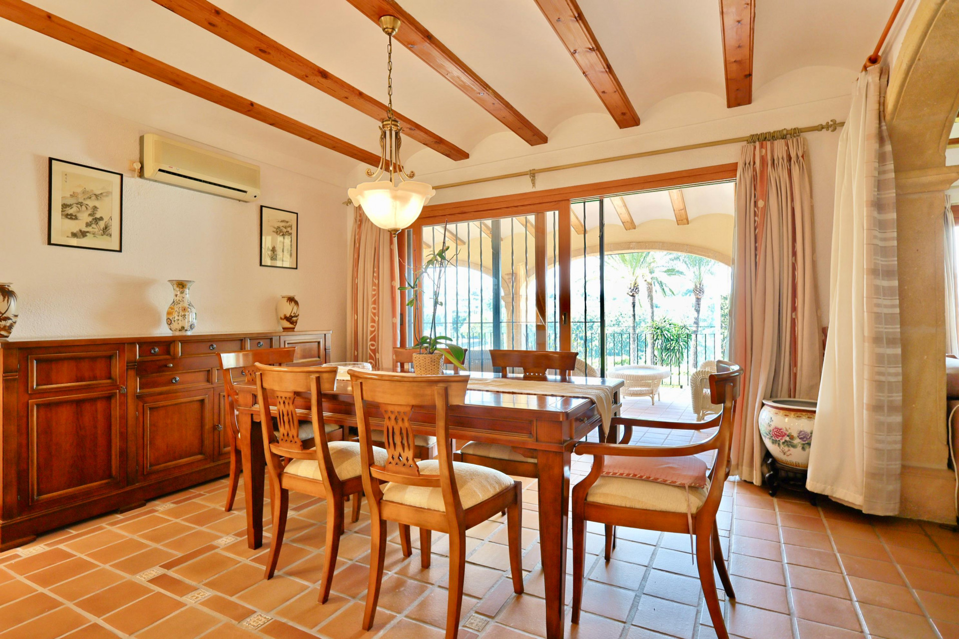 Eetkamer met houten plafondbalken, terracotta vloer en klassiek dressoir. Grote glazen schuifdeuren bieden toegang tot een overdekt terras met uitzicht.