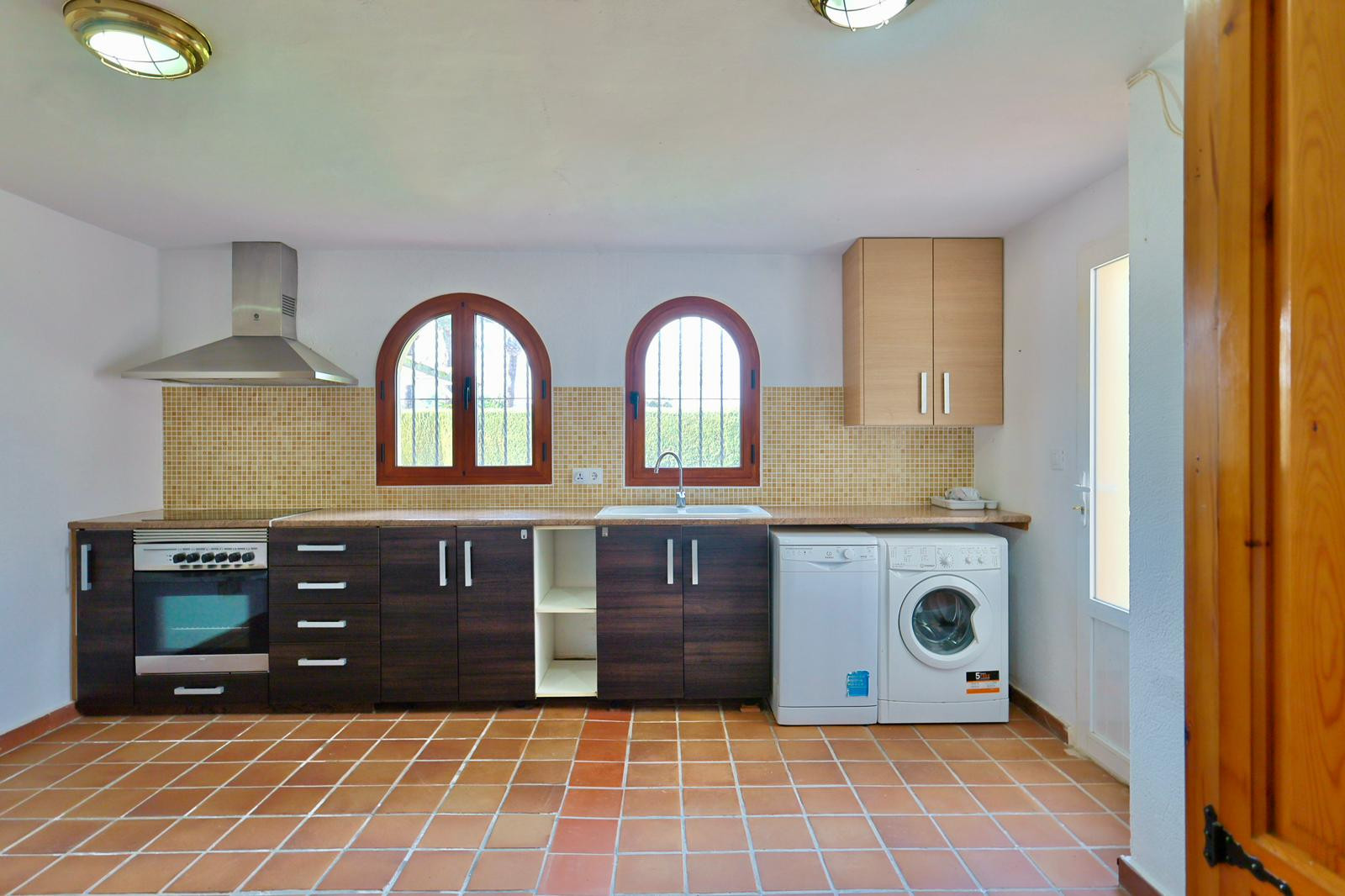 Functionele keuken en wasruimte met donkere kasten, terracotta vloer en boogvensters. Voorzien van inbouwapparatuur en een mozaïek achterwand.