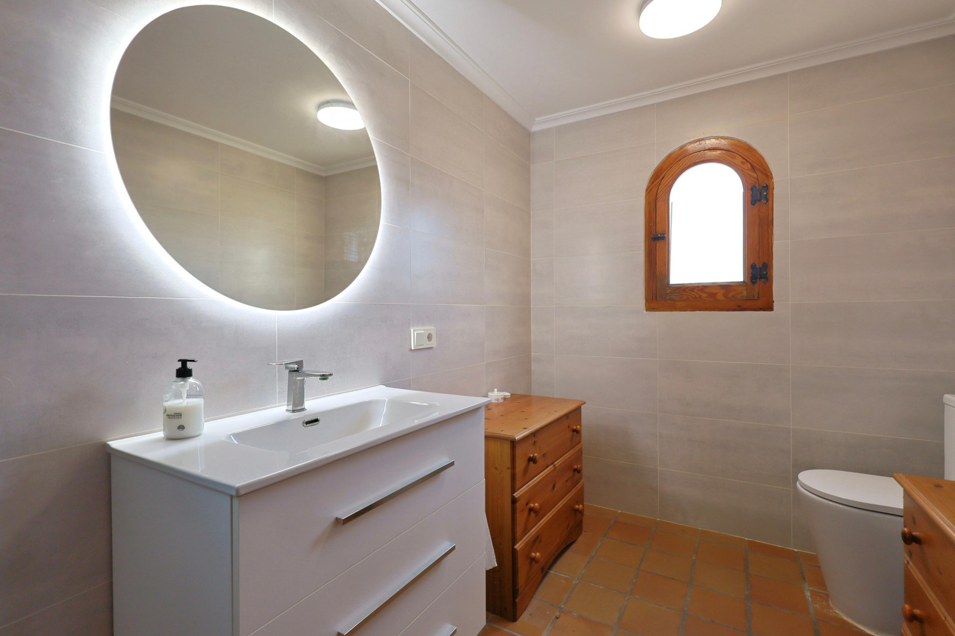 Moderne badkamer met een verlichte ronde spiegel, wit wastafelmeubel, terracotta vloertegels en een karakteristiek houten boograam.