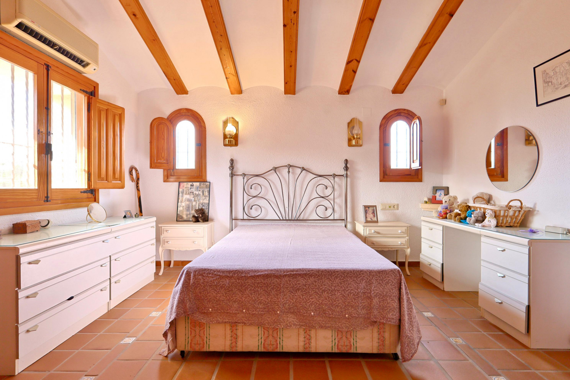 Slaapkamer met houten plafondbalken, terracotta vloertegels en boogramen met houten luiken. Voorzien van een smeedijzeren bed en split-airconditioning.