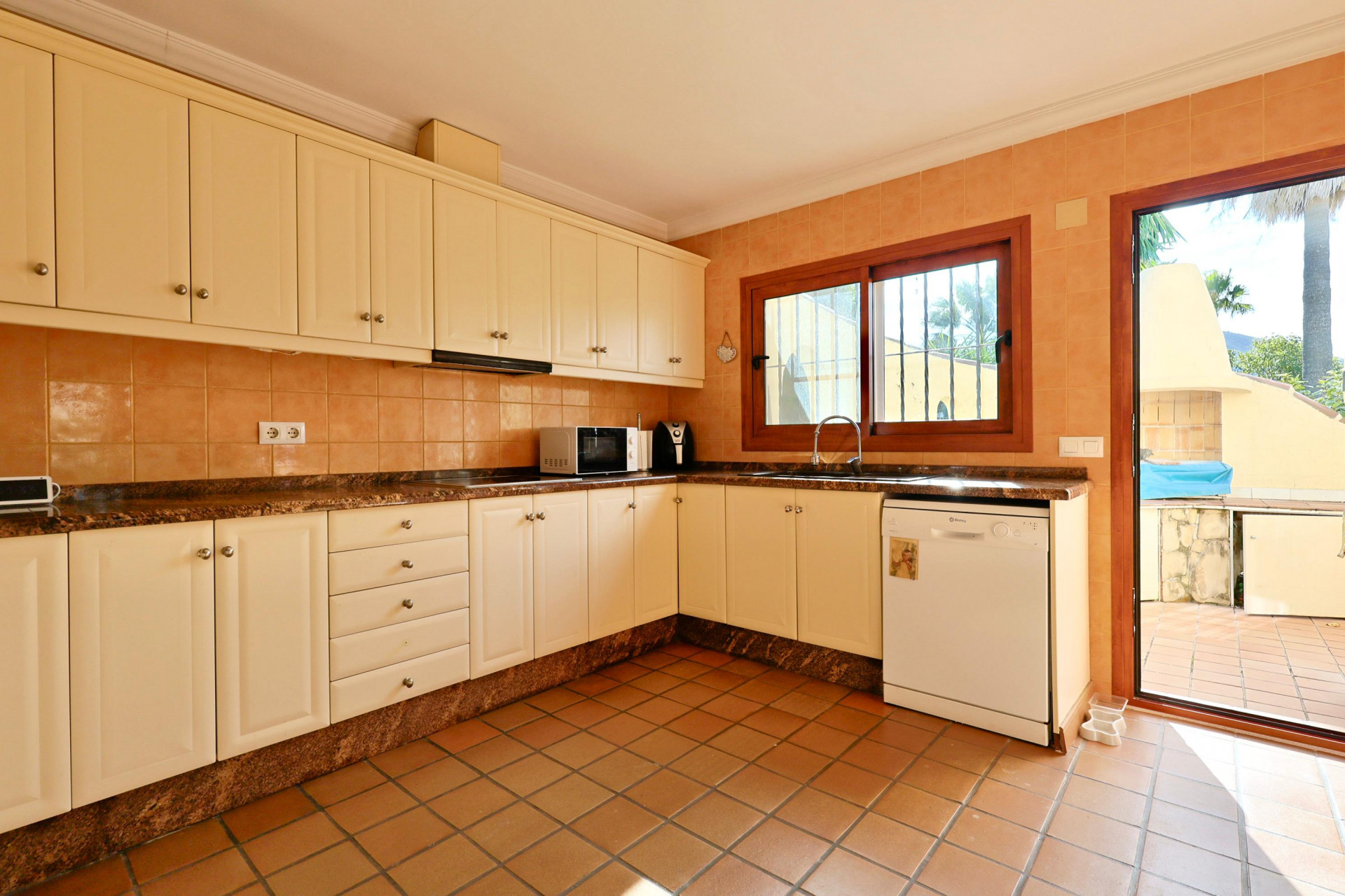 Volledig uitgeruste keuken met crèmekleurige kasten, granieten werkblad en terracotta vloer. Biedt directe toegang tot het terras en een houten raamkozijn.
