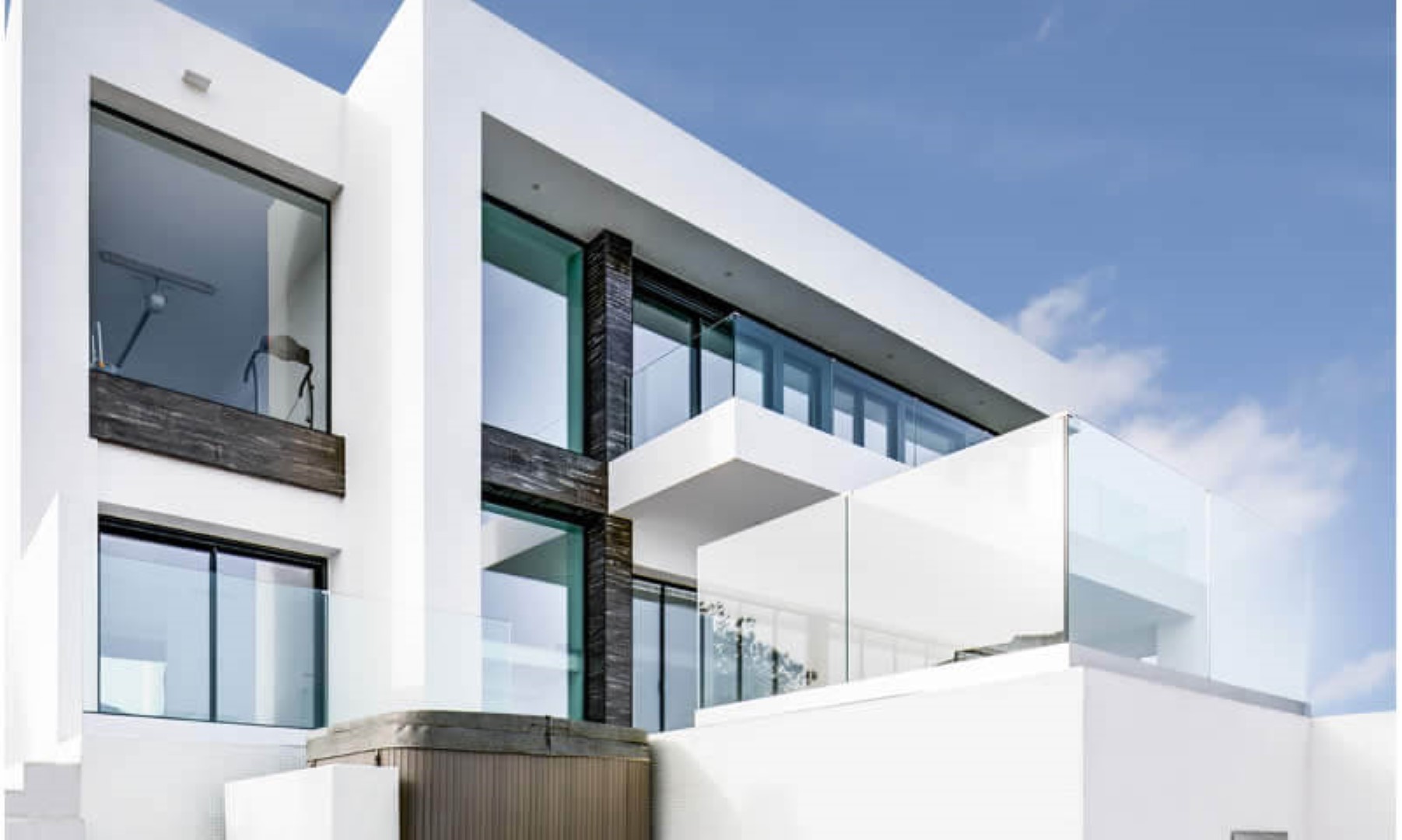 Moderne geometrische gevel met witte muren, grote raampartijen en glazen balkons. Donkere textuuraccenten contrasteren met het strakke ontwerp en de buitenjacuzzi.