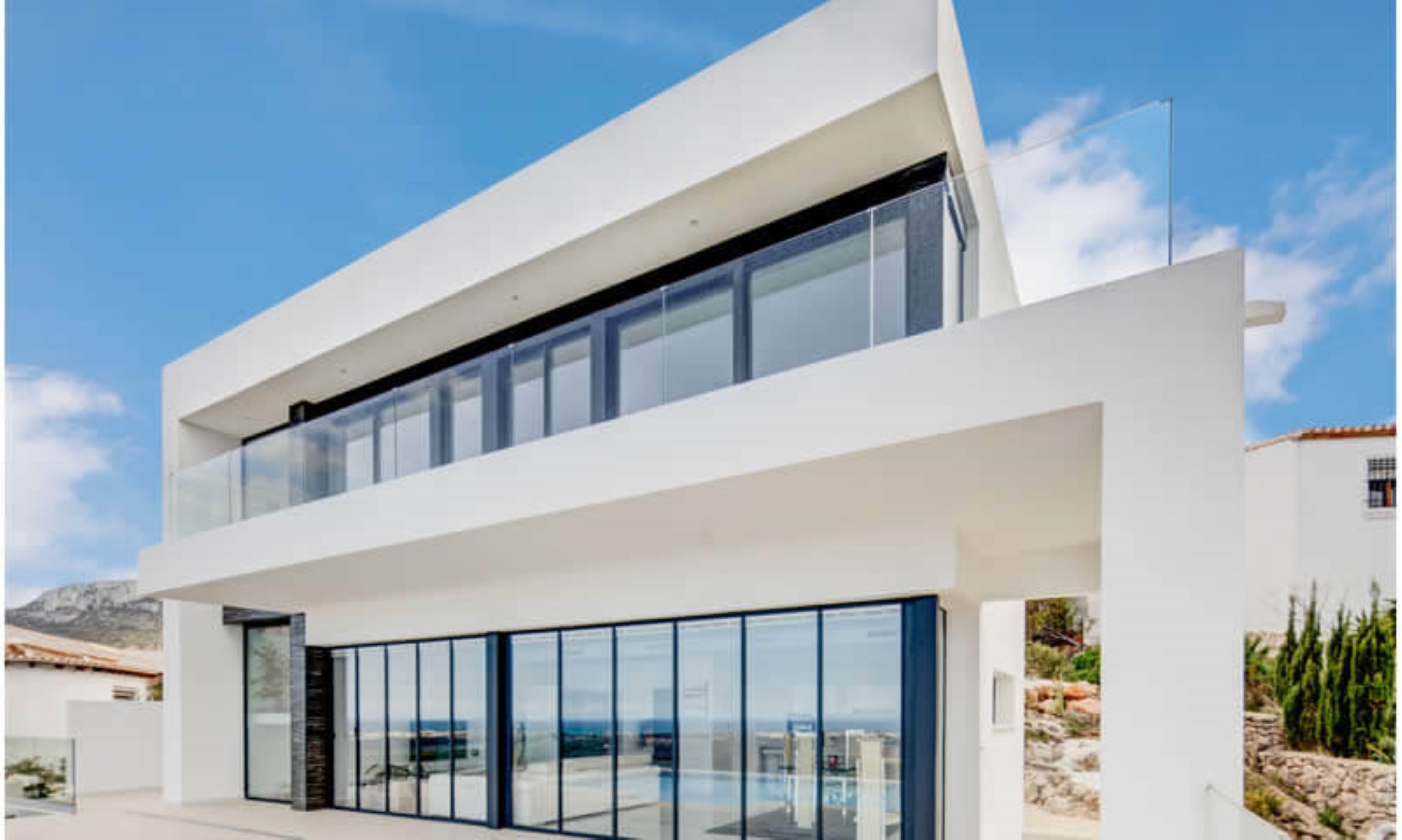 Moderne villa met twee verdiepingen, grote glazen puien van vloer tot plafond, donkere kozijnen en terrassen met glazen balustrades.
