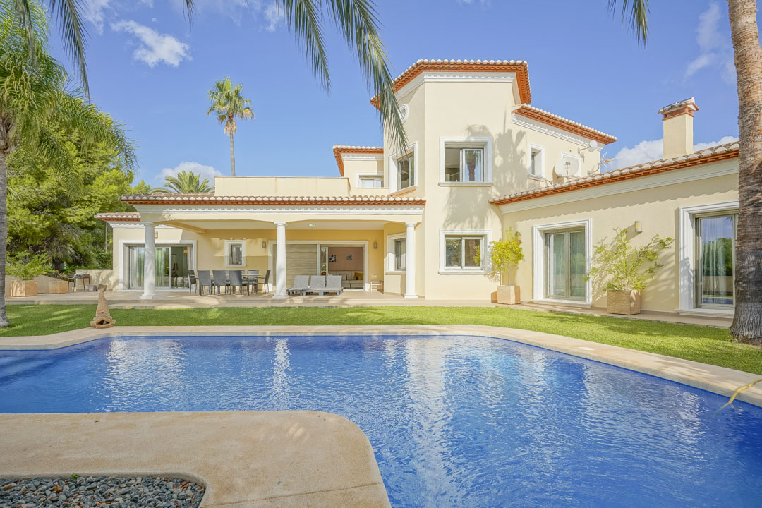 Exterieur van de villa met een groot zwembad, overdekt terras met witte kolommen en een volgroeide mediterrane tuin met palmbomen.