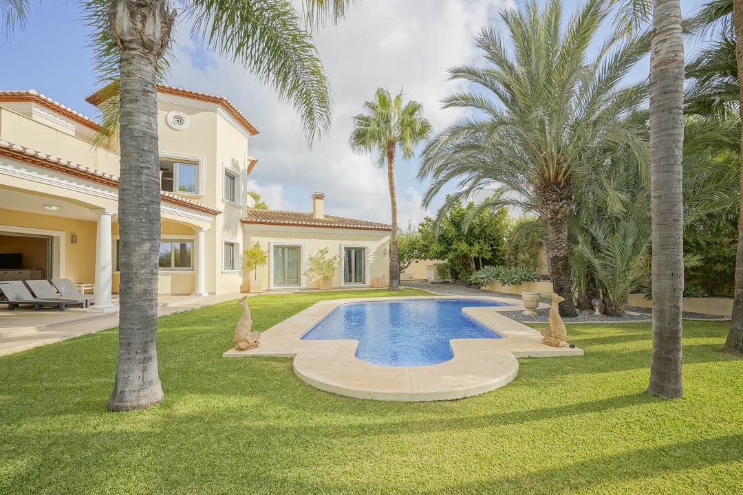 Grillig gevormd zwembad omgeven door een gazon en volgroeide palmbomen, grenzend aan de mediterrane villa met overdekt terras en witte zuilen.